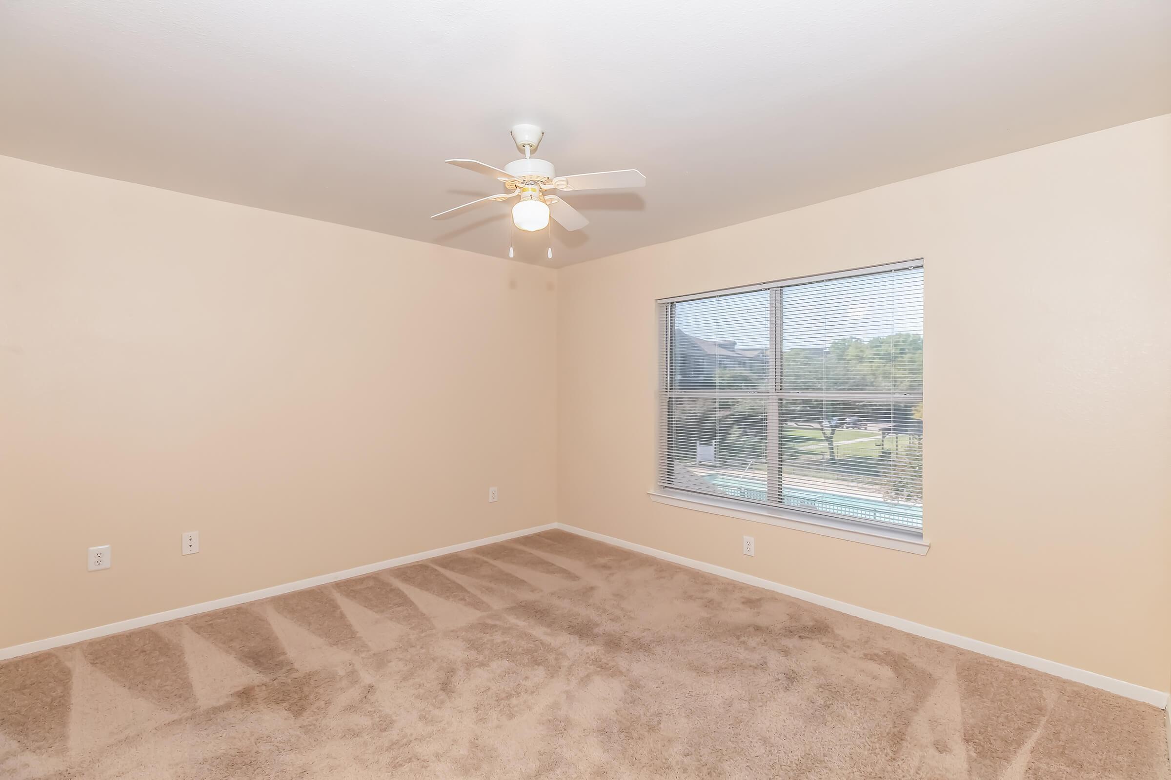 A spacious, empty bedroom featuring light beige walls and carpet. A ceiling fan is mounted in the center of the ceiling, and a large window with blinds allows natural light to enter, offering a view of outdoor greenery. The room has no furniture, creating an open and inviting atmosphere.