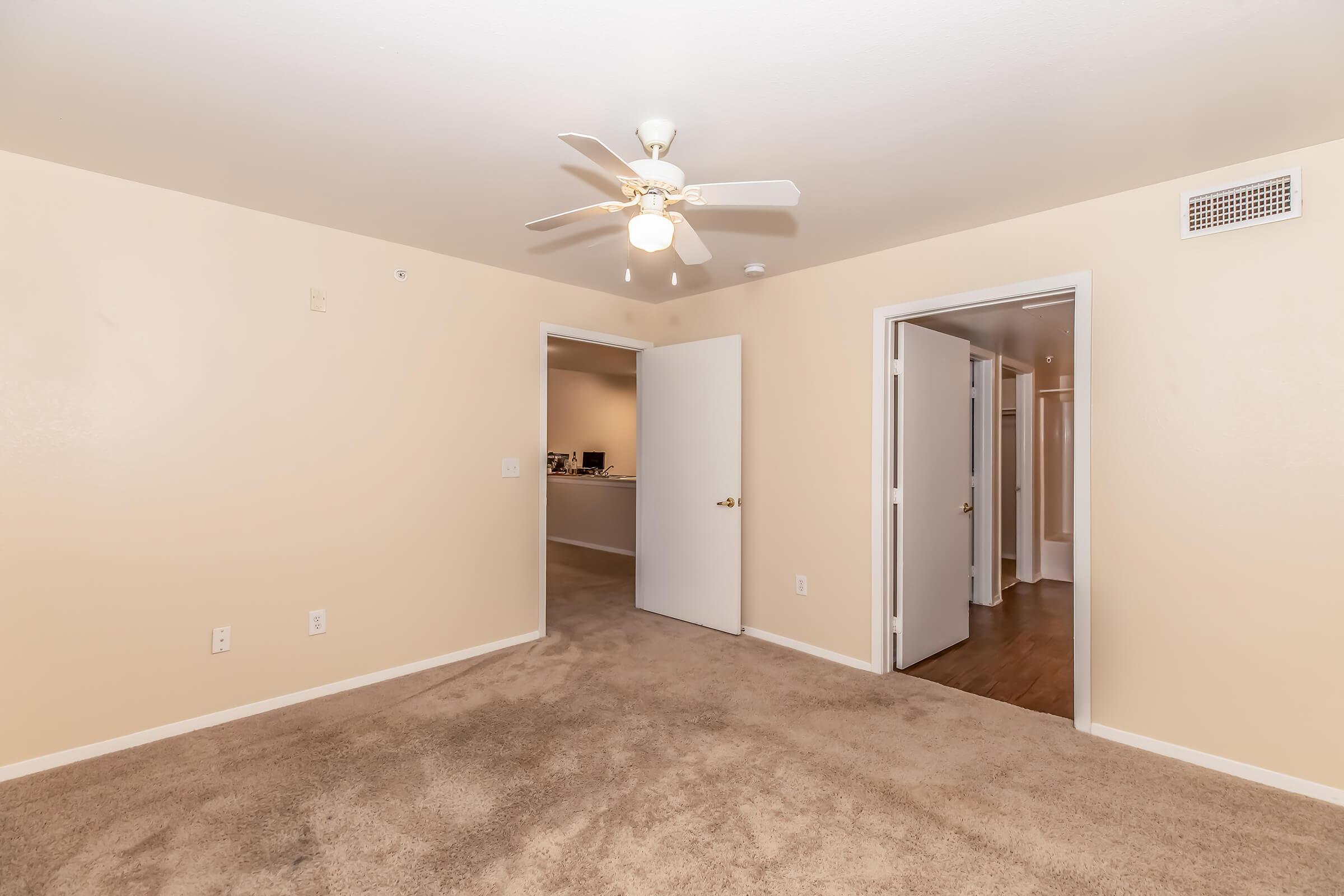 A spacious, empty room with beige walls and light-colored carpet. A ceiling fan is visible, and there are two open doors leading to adjacent spaces. The room is well-lit, creating a clean and inviting atmosphere, suitable for various uses like a living area or bedroom.