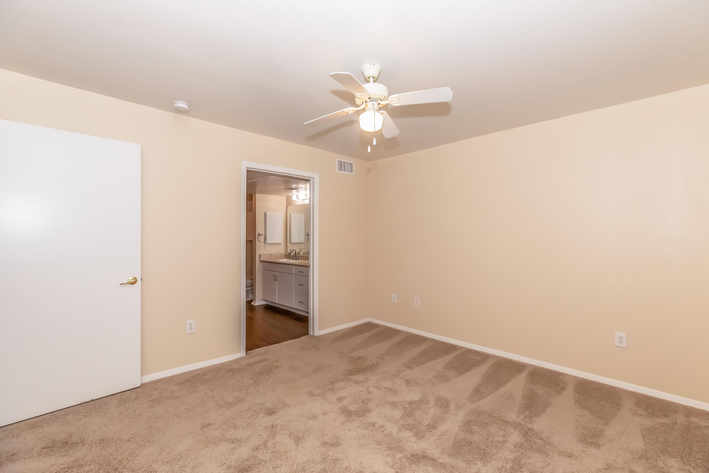 A spacious, empty room featuring beige walls and a light brown carpet. There is a ceiling fan with five blades and a white door on the left. An open doorway leads to a bathroom, visible in the background with bright lighting and a mirror. The room is clean and well-maintained, ideal for various uses.