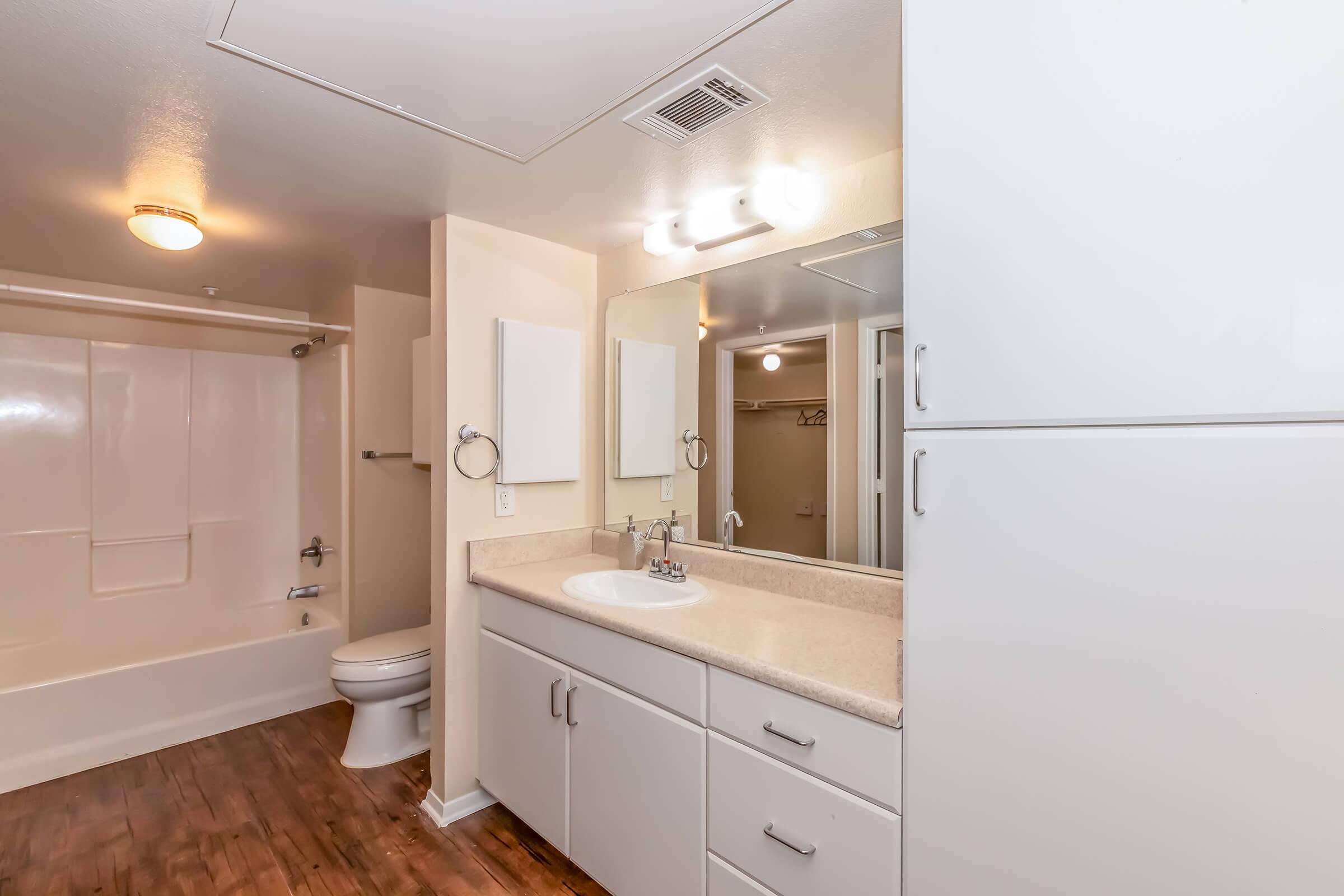 A modern bathroom featuring a white vanity with a sink, a large mirror, and a light fixture overhead. The room includes a white toilet, a bathtub with a shower, and light wood flooring. The walls are painted in a neutral color, creating a spacious and bright atmosphere.