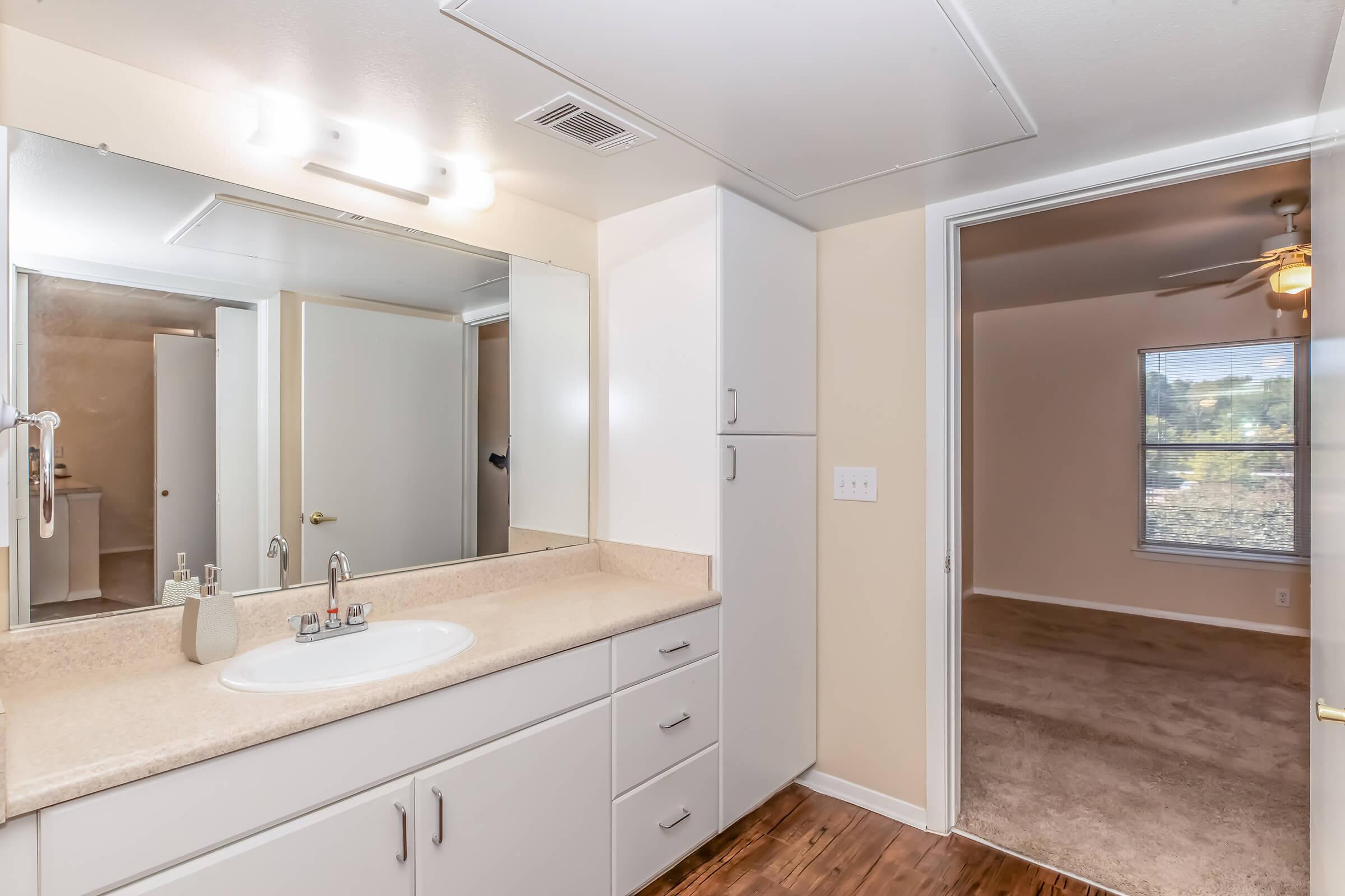 Bright bathroom featuring a large mirror above a beige countertop with a sink and storage drawers. A door leads to a carpeted room with natural light streaming in through a window. The walls are painted in a neutral tone, creating an airy and spacious atmosphere.