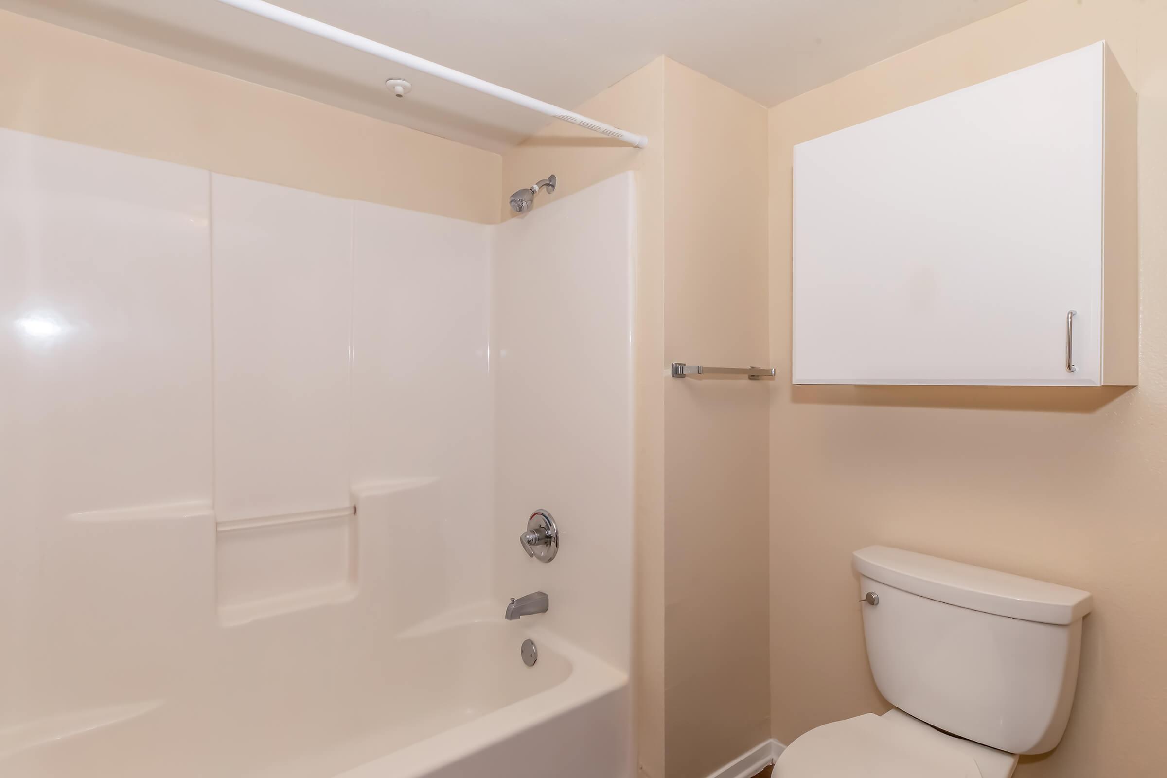 A clean and simple bathroom featuring a white bathtub with a showerhead, a toilet, and a white cabinet mounted on the wall. The walls are painted in a light beige color, creating a bright and airy atmosphere.