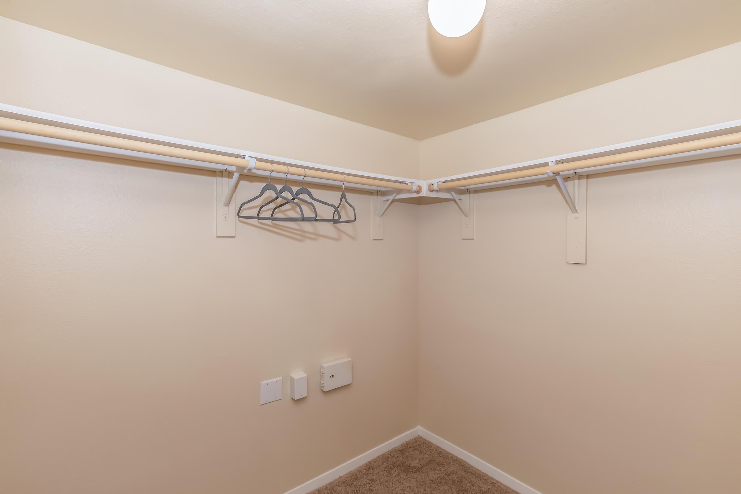 An empty walk-in closet featuring two hanging rods with several empty hangers. The walls are painted in a light beige color, and there are electrical outlets on one side. The floor is covered with a light brown carpet, and a ceiling light fixture provides illumination.