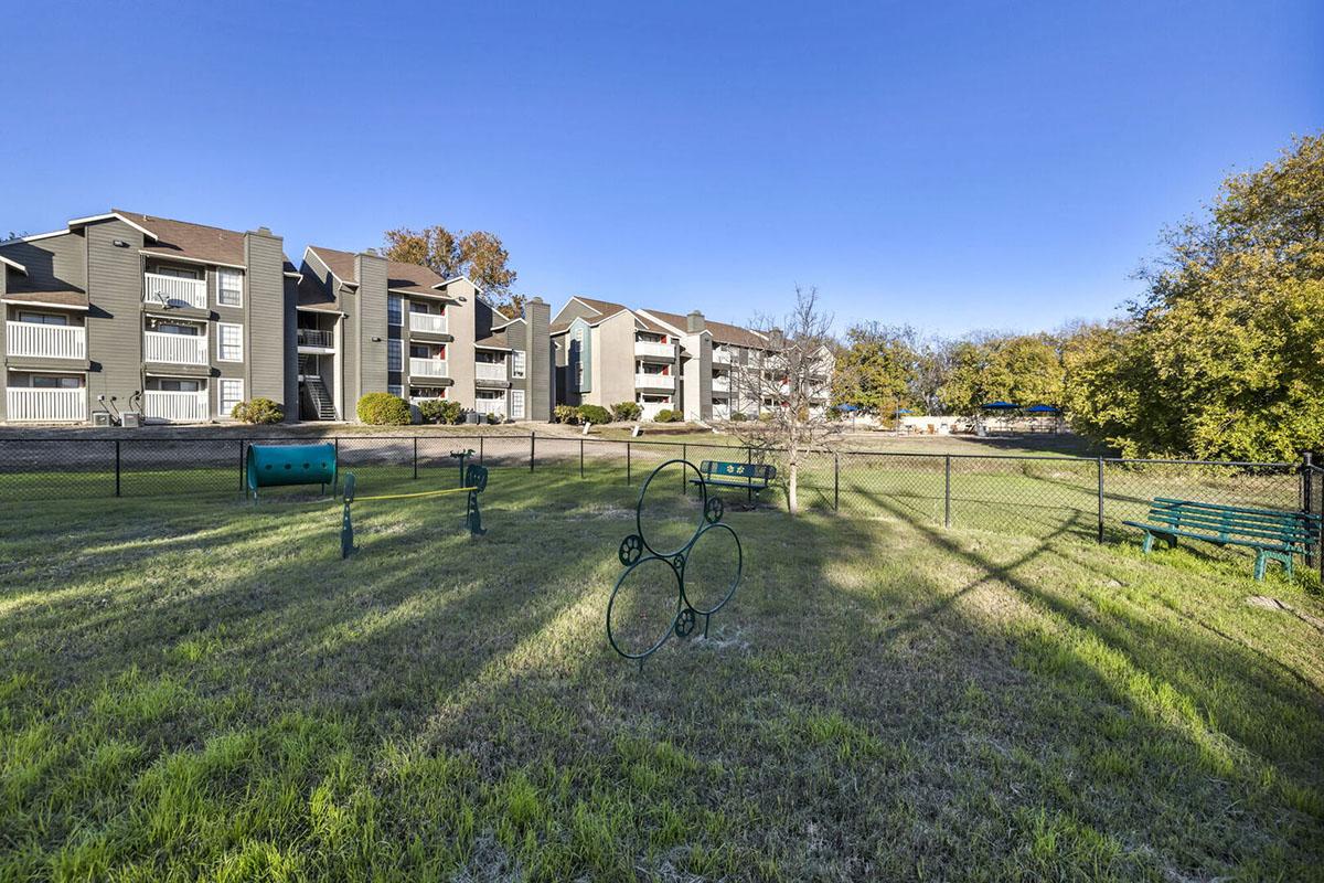A well-maintained grassy area with exercise equipment, surrounded by a fence. In the background, there are several multi-story apartment buildings with balconies. The scene is set under a clear blue sky, with trees providing some shade.