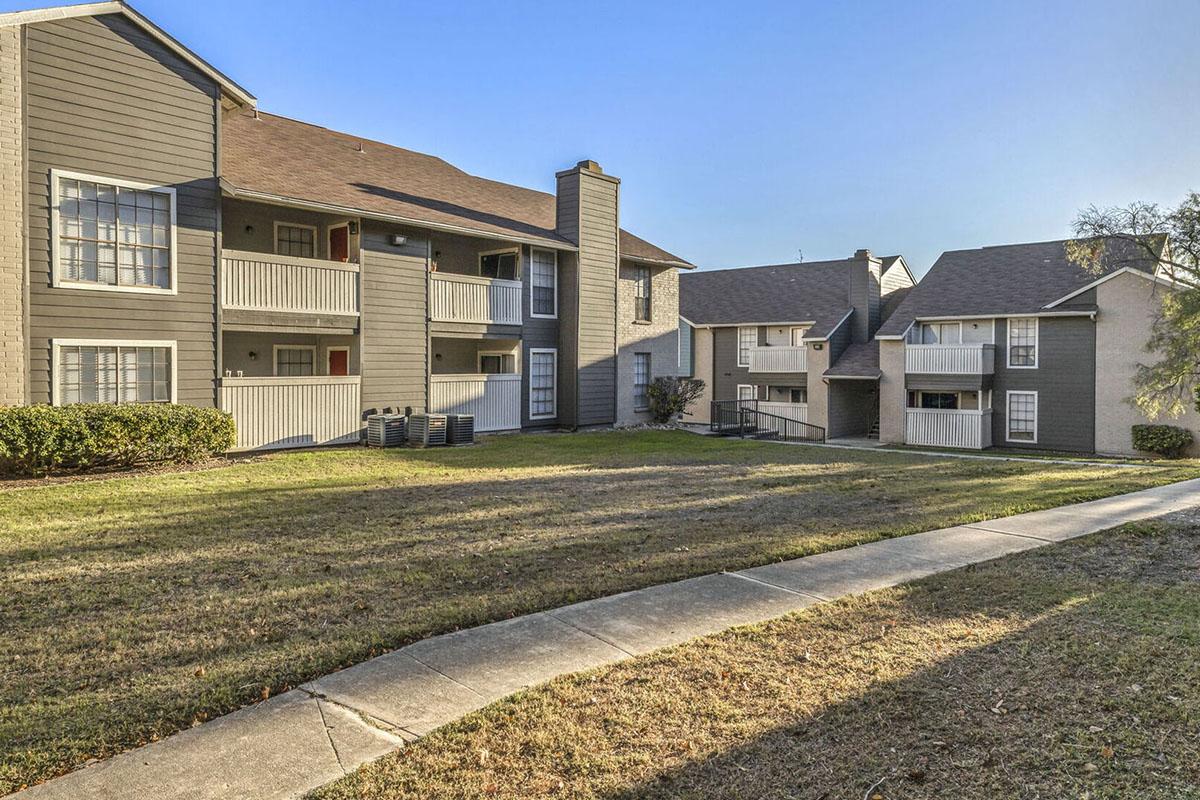 A view of a two-story apartment complex with multiple units. The buildings feature balconies and are surrounded by grassy lawns. A concrete path leads through the landscape, connecting the buildings. The sky is clear, indicating a sunny day.
