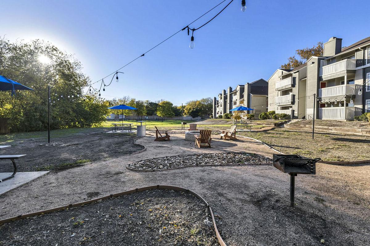 A landscaped outdoor area featuring a fire pit with seating, picnic tables, and blue umbrellas. String lights are hung overhead, with green grassy spaces and modern apartment buildings in the background under a clear blue sky.