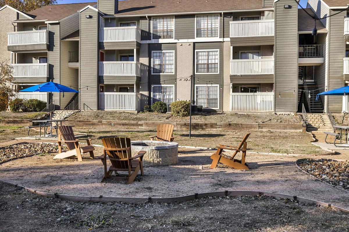 Outdoor area featuring a fire pit surrounded by four wooden Adirondack chairs, set within a landscaped space. In the background, there are two-story residential buildings with balconies, and two blue umbrellas providing shade near picnic tables. The ground is covered with gravel and grass.