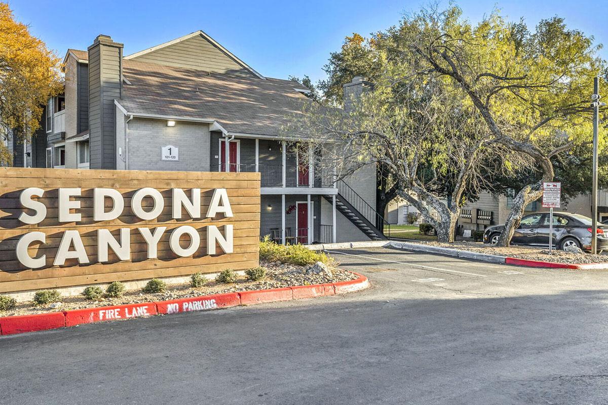Apartment complex with a sign reading "Sedona Canyon." The building features two stories with a mix of gray and brown siding, surrounded by trees with autumn foliage. A parking lot is visible, and there are red-painted curbs indicating a fire lane.