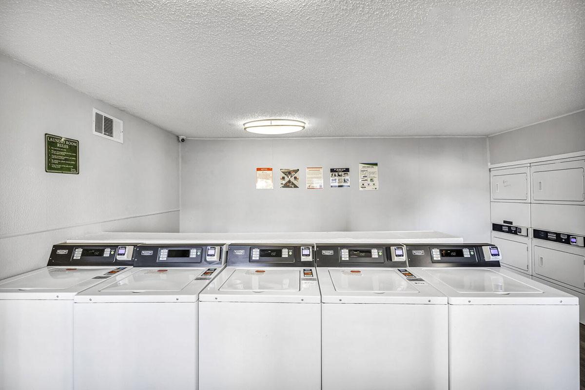 A clean and well-lit laundry room featuring four white washing machines aligned in a row. The walls are painted in light gray, with a ceiling light fixture providing illumination. Informational posters are visible, along with a set of storage cabinets to the side.