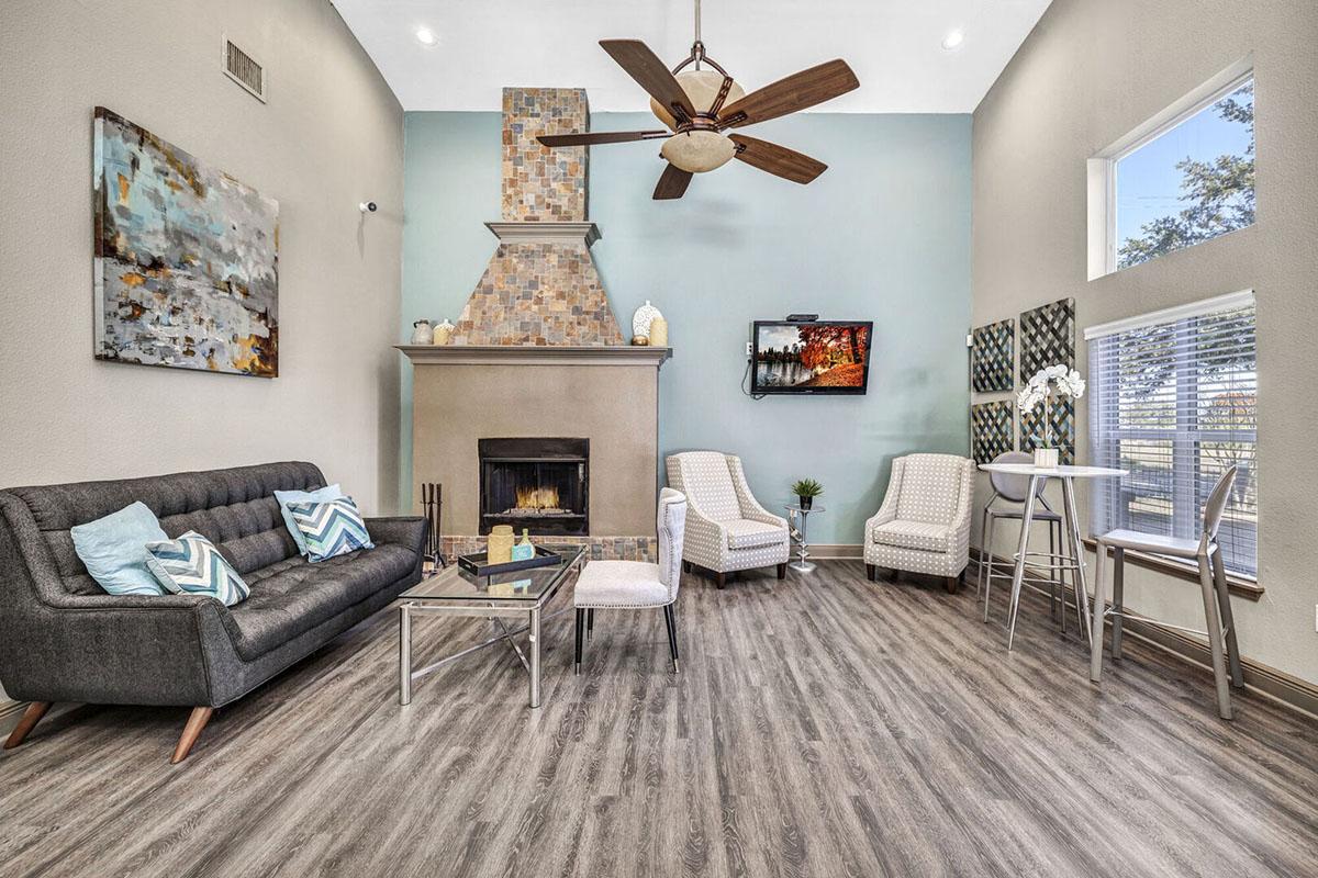 A modern living room featuring a gray couch with decorative pillows, two wicker chairs, a coffee table, and a fireplace with a tiled surround. The walls are painted light blue and beige, and there is a mounted TV with artwork on the walls. Natural light floods the space through large windows.