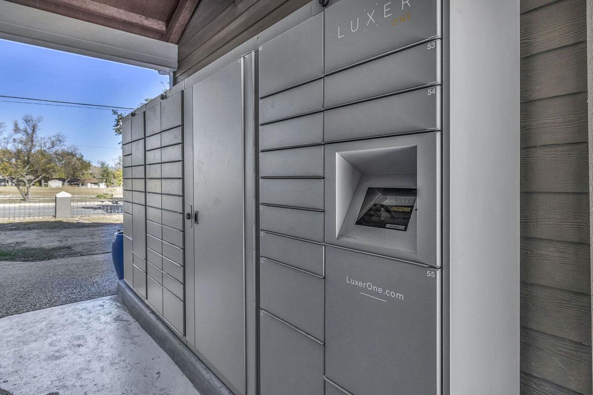 A modern outdoor delivery locker system with various compartments for storing packages, located next to a house. The locker includes a drop-off slot and features the logo "LUXER ONE." The surrounding area shows a clear sky and a nearby fence.