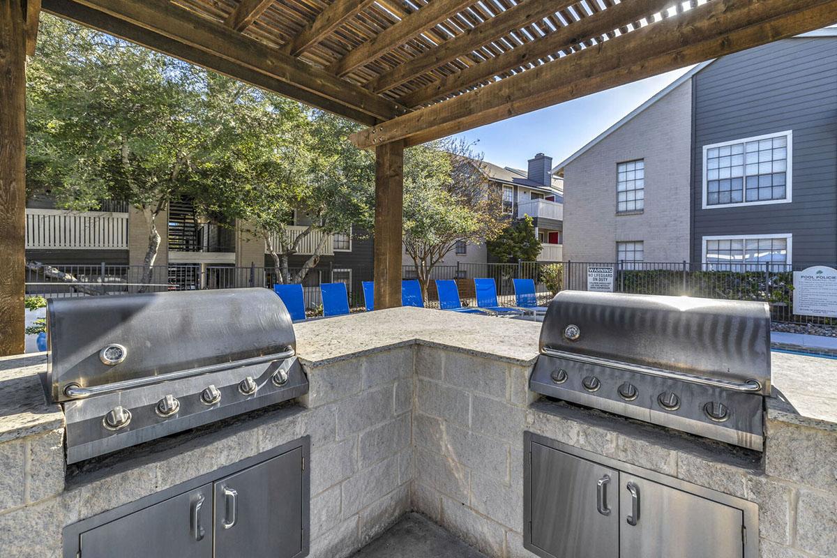 Outdoor grilling area featuring two stainless steel barbecue grills and a stone countertop. In the background, there is a swimming pool surrounded by blue lounge chairs and residential buildings with large windows. The space is shaded by a wooden pergola and lush trees.