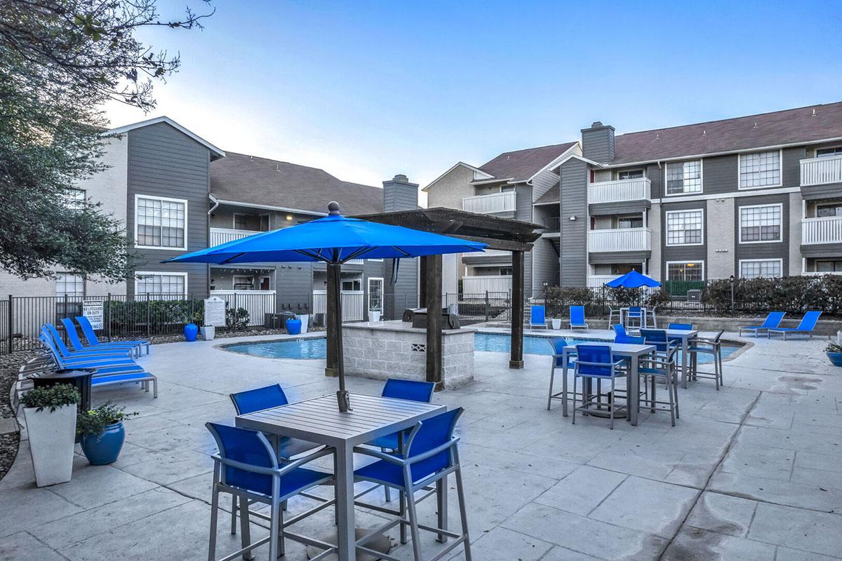 A tranquil apartment complex pool area featuring a blue umbrella, lounge chairs, and dining tables. The pool is surrounded by modern buildings with balconies, and landscaped greenery adds a touch of nature to the scene. The setting is inviting and ideal for relaxation or social gatherings.