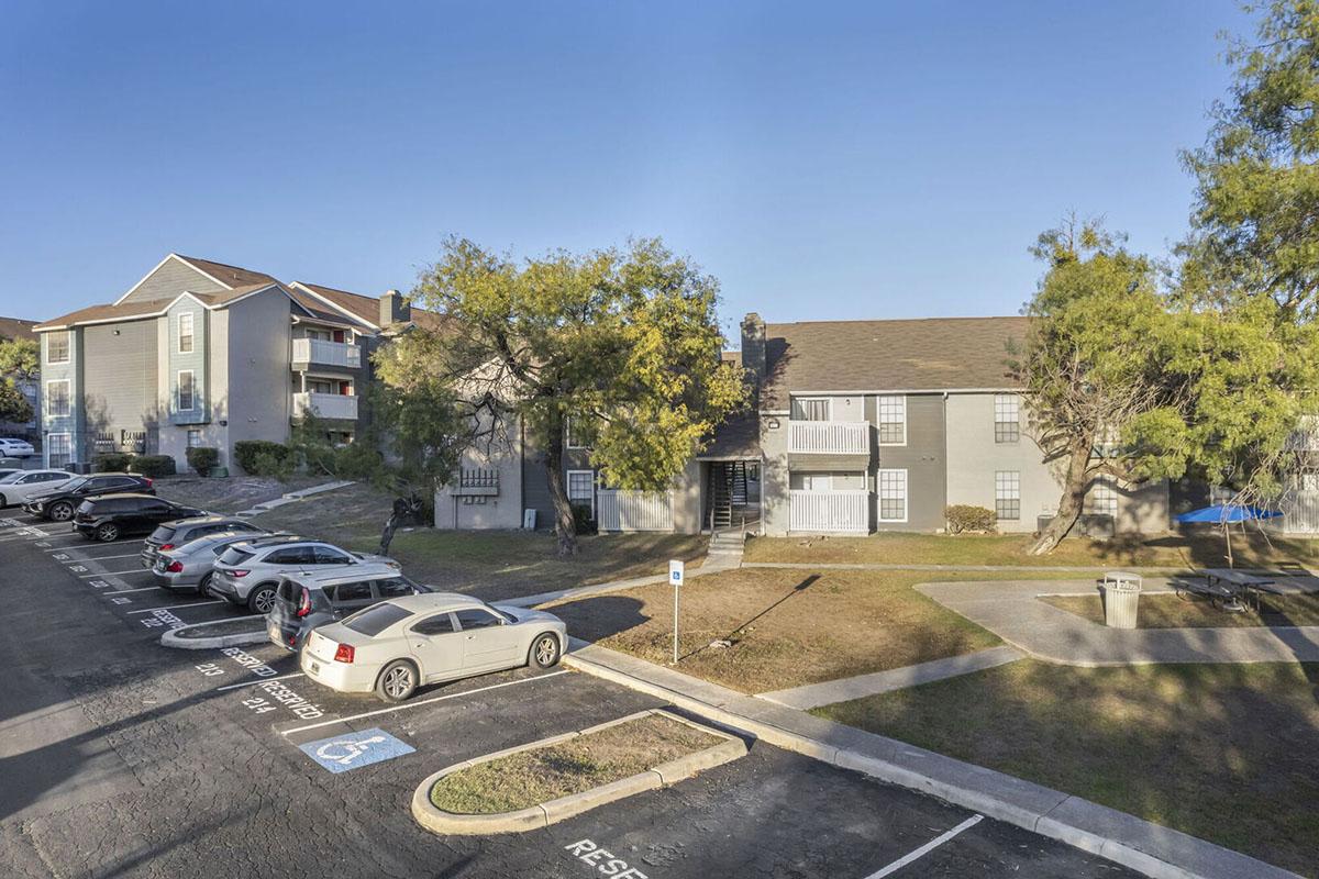 A view of an apartment complex with two buildings, one featuring multiple windows and a balcony. The foreground includes a parking lot with several cars, including designated handicapped spots. Surrounding the buildings are trees and a grassy area, under a clear blue sky.