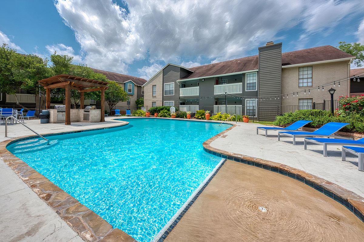 A vibrant swimming pool surrounded by lounge chairs and greenery. In the background, there are two-story apartment buildings with balconies. The sky is partly cloudy, creating a sunny and inviting atmosphere. A pergola and flower beds enhance the outdoor setting.