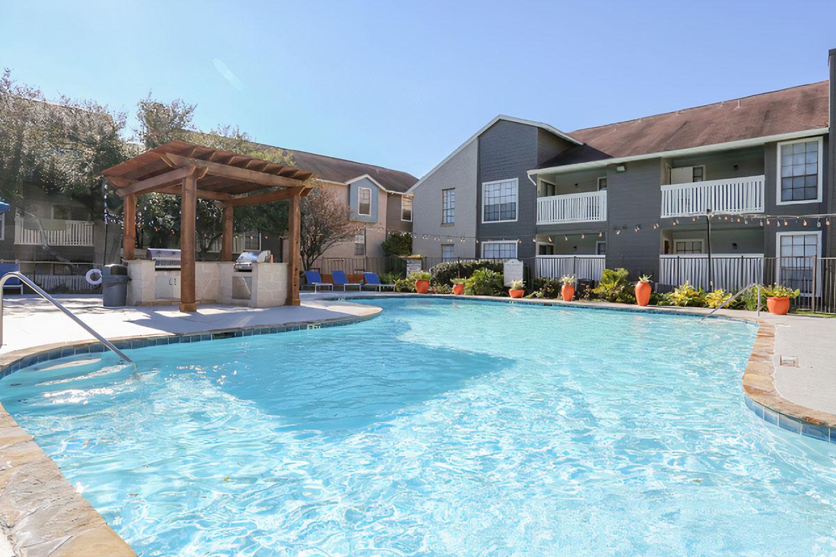 A clear blue swimming pool with a stone border, surrounded by lounge chairs and potted plants. In the background, there are two multi-story buildings with balconies. A pergola with outdoor cooking facilities is located near the pool, set against a sunny sky.