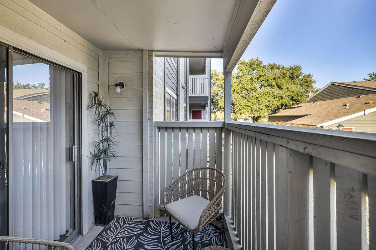 A small balcony with a wicker chair and a potted plant. The area features light wood paneling and a patterned outdoor rug. There's a view of neighboring buildings, and the sky is clear. The balcony has a railing and sliding glass doors, creating a cozy outdoor space.