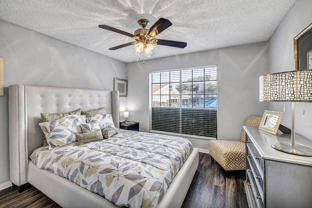 A cozy bedroom featuring a queen-sized bed with a decorative headboard, patterned bedding, a stylish ceiling fan, and a large window allowing natural light. The room includes a bedside table with a lamp, a decorative chair, and a dresser, creating a comfortable and inviting atmosphere.