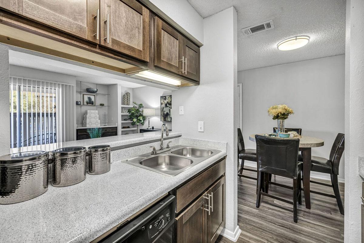 Modern kitchen interior with a double sink and stainless steel appliances. The countertop is light-colored with decorative storage containers. In the background, an open living space features a dining table with four chairs, a vase with flowers, and a shelf displaying home decor.