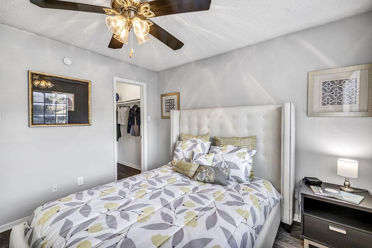 A cozy bedroom featuring a queen-sized bed with a light floral comforter, a bedside table with a lamp, and a ceiling fan with lights. The walls are painted light gray, and there's a framed mirror and artwork. A door leads to a closet area, enhancing the room's functional appeal.