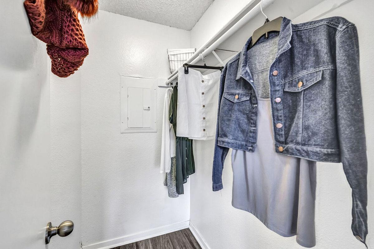 A small closet with a white wall, featuring a hanging denim jacket and several items of clothing, including a white skirt and a green top. A textured scarf is hanging on the door handle, and a wall panel is visible near the left side of the image. The floor is dark wood.
