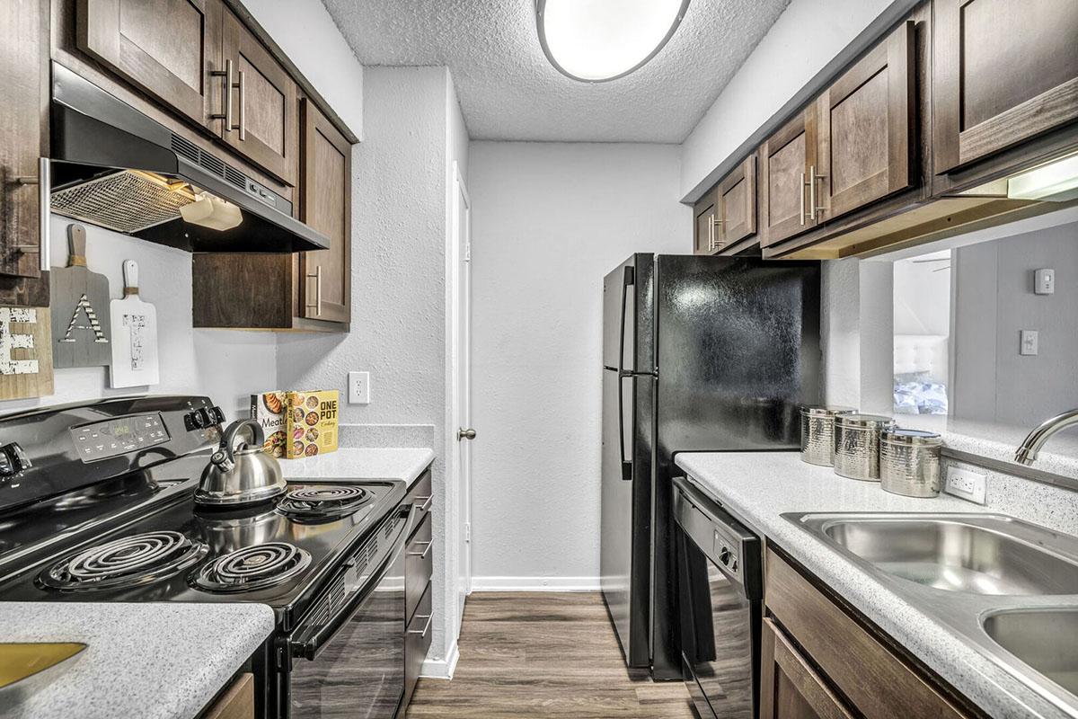 Modern kitchen featuring dark wooden cabinets, a black refrigerator, stove, and overhead microwave. Countertops are light-colored, and there are stainless steel sinks. The room has a clean, contemporary design with a window letting in natural light and various kitchen essentials on display.