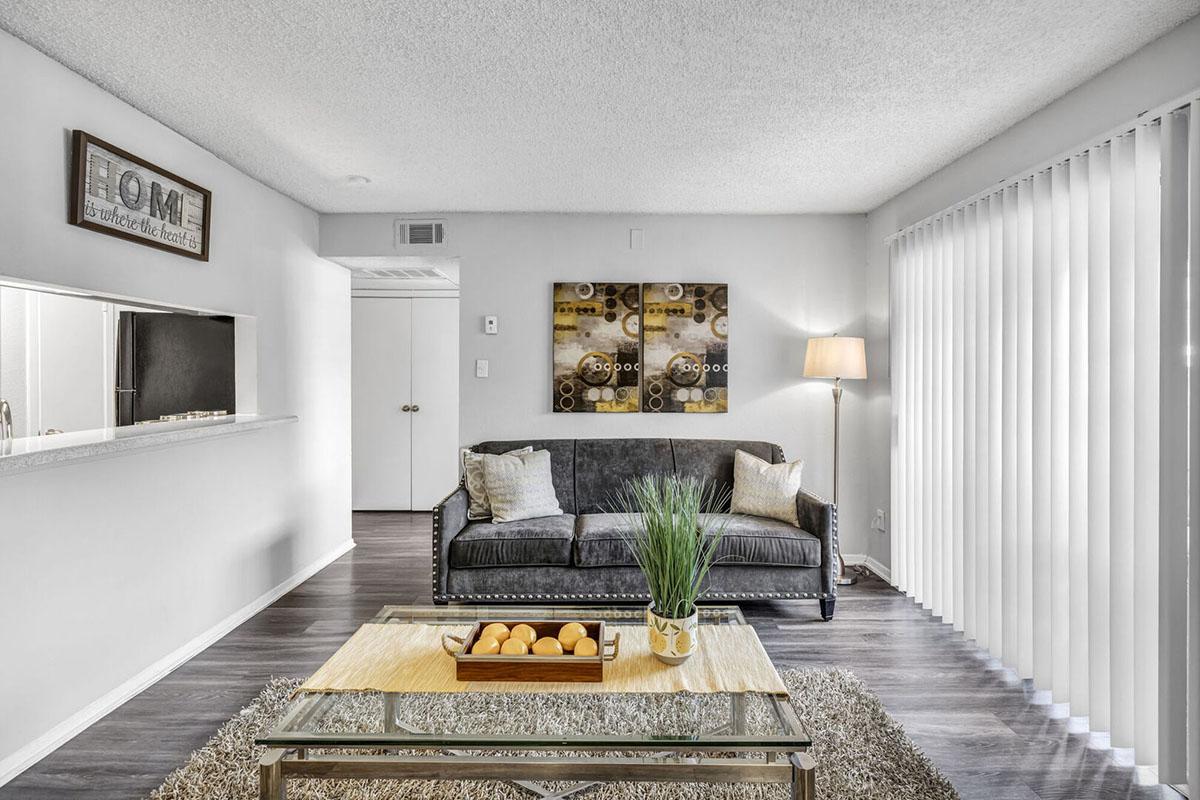 A cozy living room featuring a dark gray sofa with decorative pillows, a glass coffee table with a bowl of oranges, and a small potted plant. The space is well-lit with a floor lamp, has wood-like flooring, and large sliding glass doors with vertical blinds. Wall art decorates the wall above the sofa.