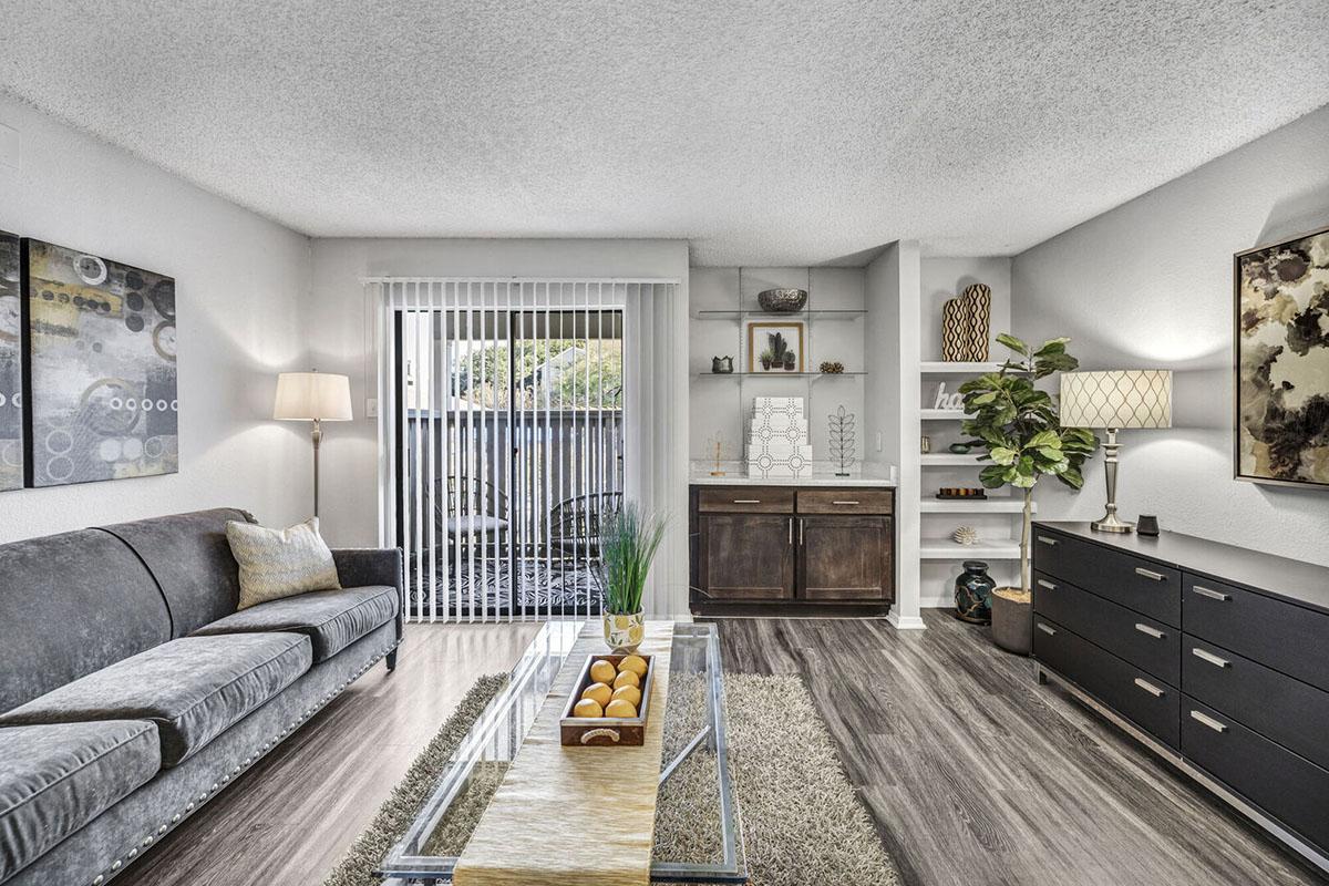 A modern living room featuring a gray sofa, glass coffee table with decorative items, a dark wooden cabinet, and tasteful wall art. The space has a light color palette, large windows with blinds, and a cozy atmosphere enhanced by a floor lamp and indoor plants.