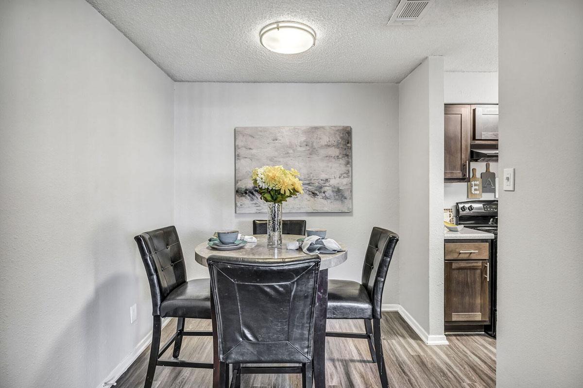 A modern dining area featuring a round table set with blue plates and a vase of yellow flowers. Four black chairs surround the table. A painting hangs on the wall, adding a decorative touch to the neutral-colored space. The kitchen is visible in the background with dark cabinetry and appliances.