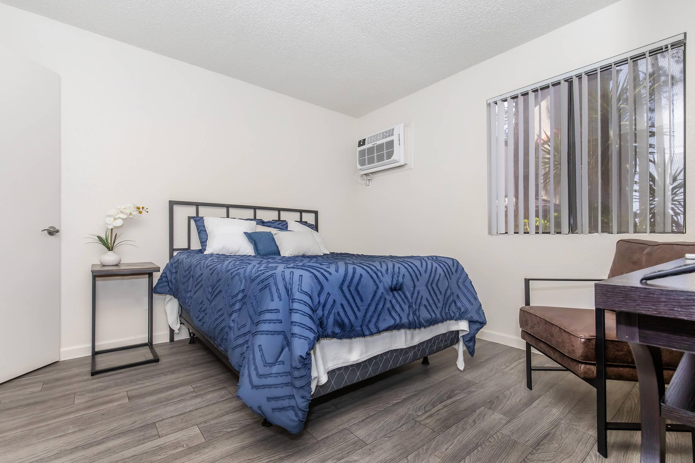 A cozy bedroom featuring a double bed with blue bedding, a nightstand with a decorative plant, a chair, and an air conditioning unit on the wall. The room has light-colored walls and a window with vertical blinds, providing natural light and a view of greenery outside.
