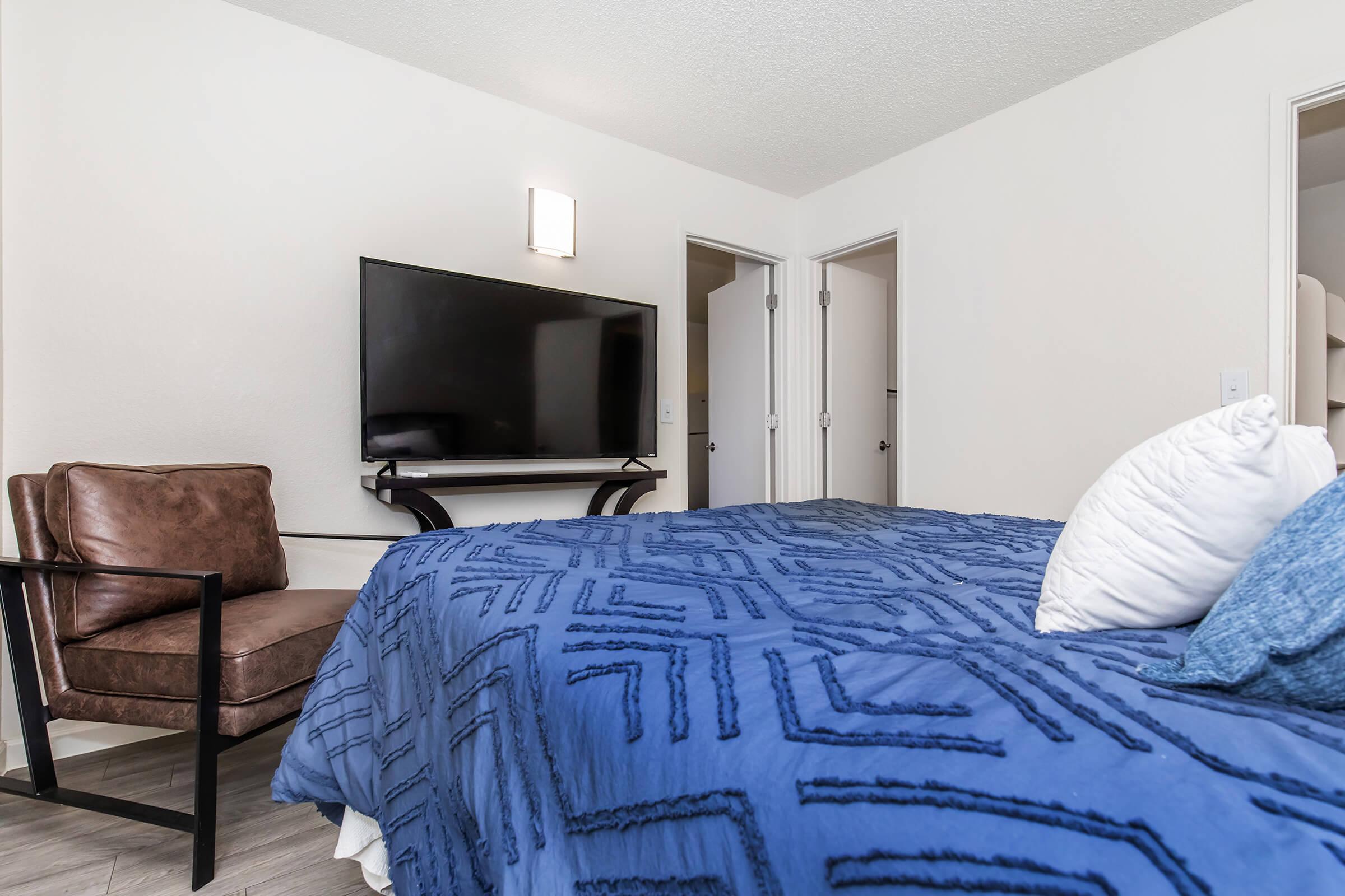 A cozy bedroom featuring a blue patterned bedspread, a large wall-mounted TV, and a comfy brown armchair. There are two closed doors in the background, indicating access to a bathroom or closet. The walls are light-colored, creating a bright and inviting atmosphere.