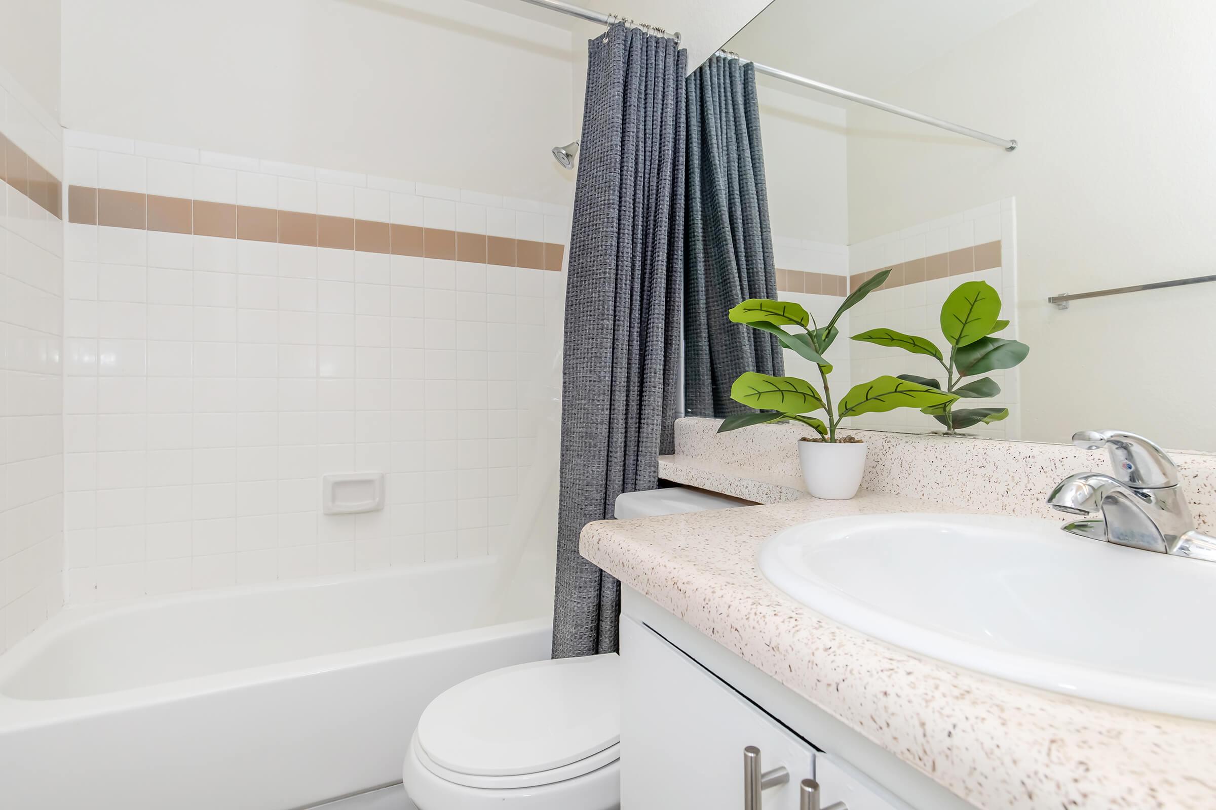 A modern bathroom featuring a white bathtub with a shower curtain, a light-colored countertop with a sink, and a small potted plant. The decor is simple and minimalistic, with beige and white tiled walls, providing a clean and bright atmosphere.