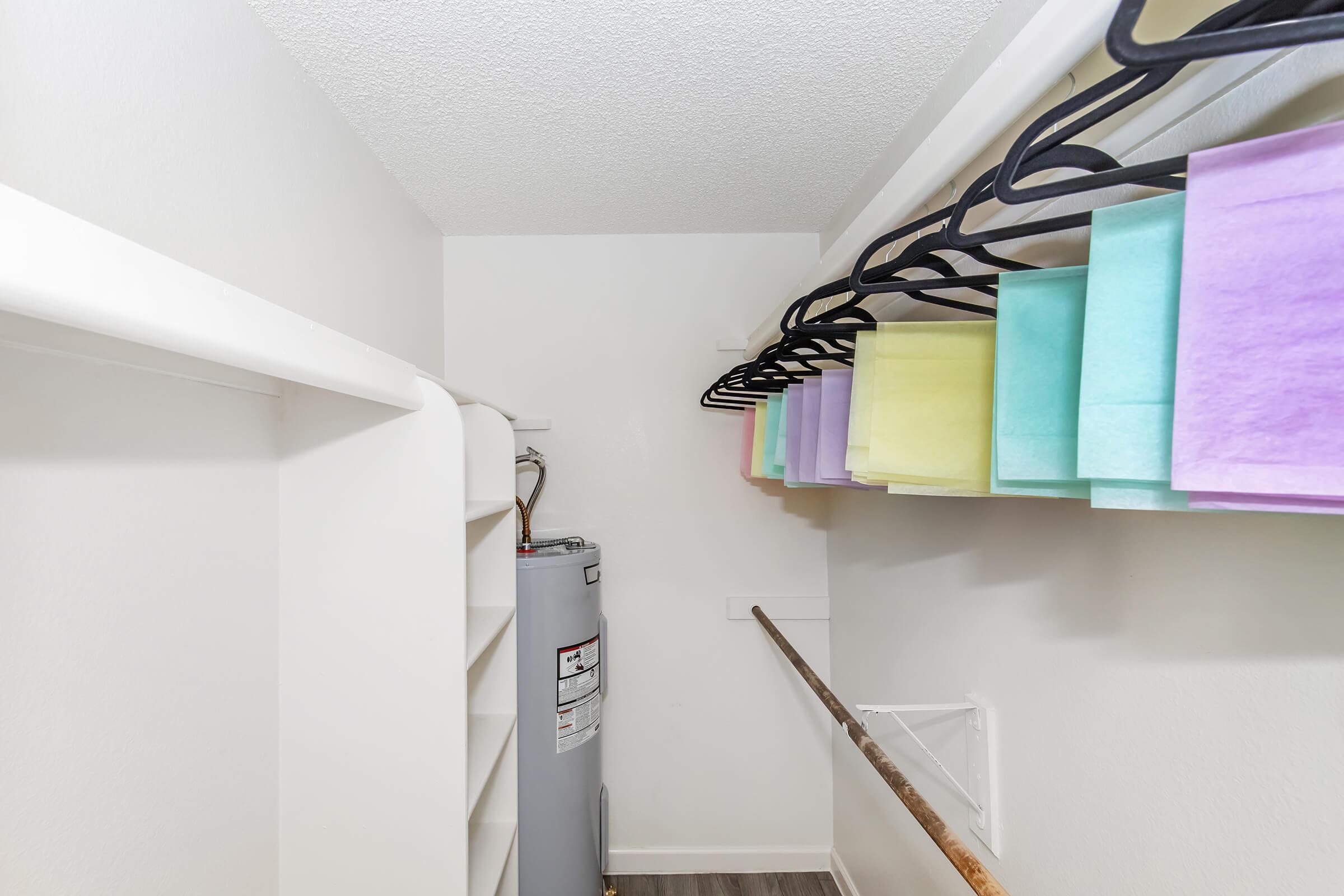 A small, well-lit closet featuring a water heater on the left and a hanging rod with pastel-colored paper sheets. The closet has white walls and a light-colored floor, with shelving along one side for additional storage.
