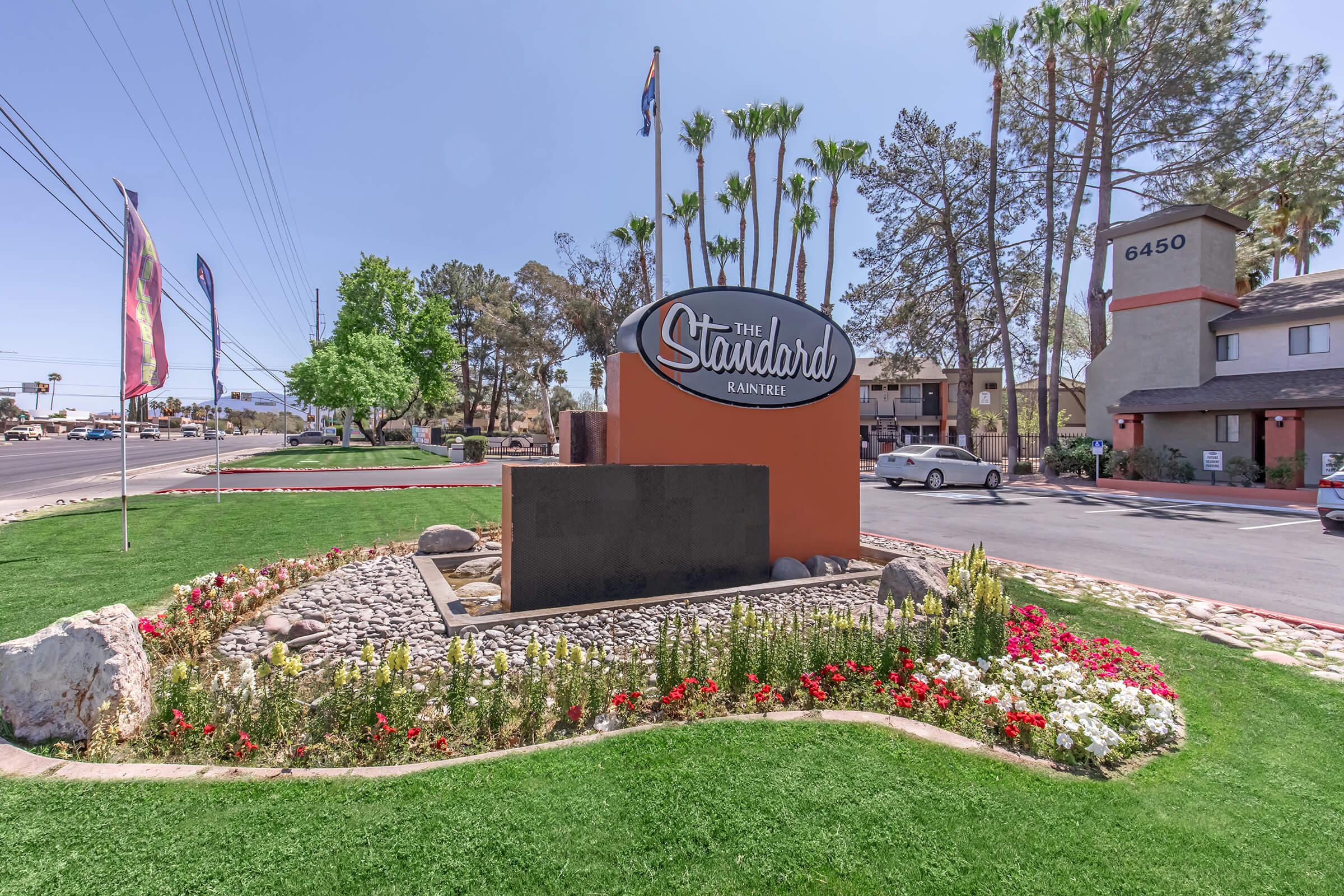 Sign for The Standard Rainier hotel, surrounded by landscaped greenery, flowers, and decorative rocks. The hotel building is visible in the background along with palm trees, and there are flags flying nearby. The setting appears sunny and inviting, typical of a hospitality location.