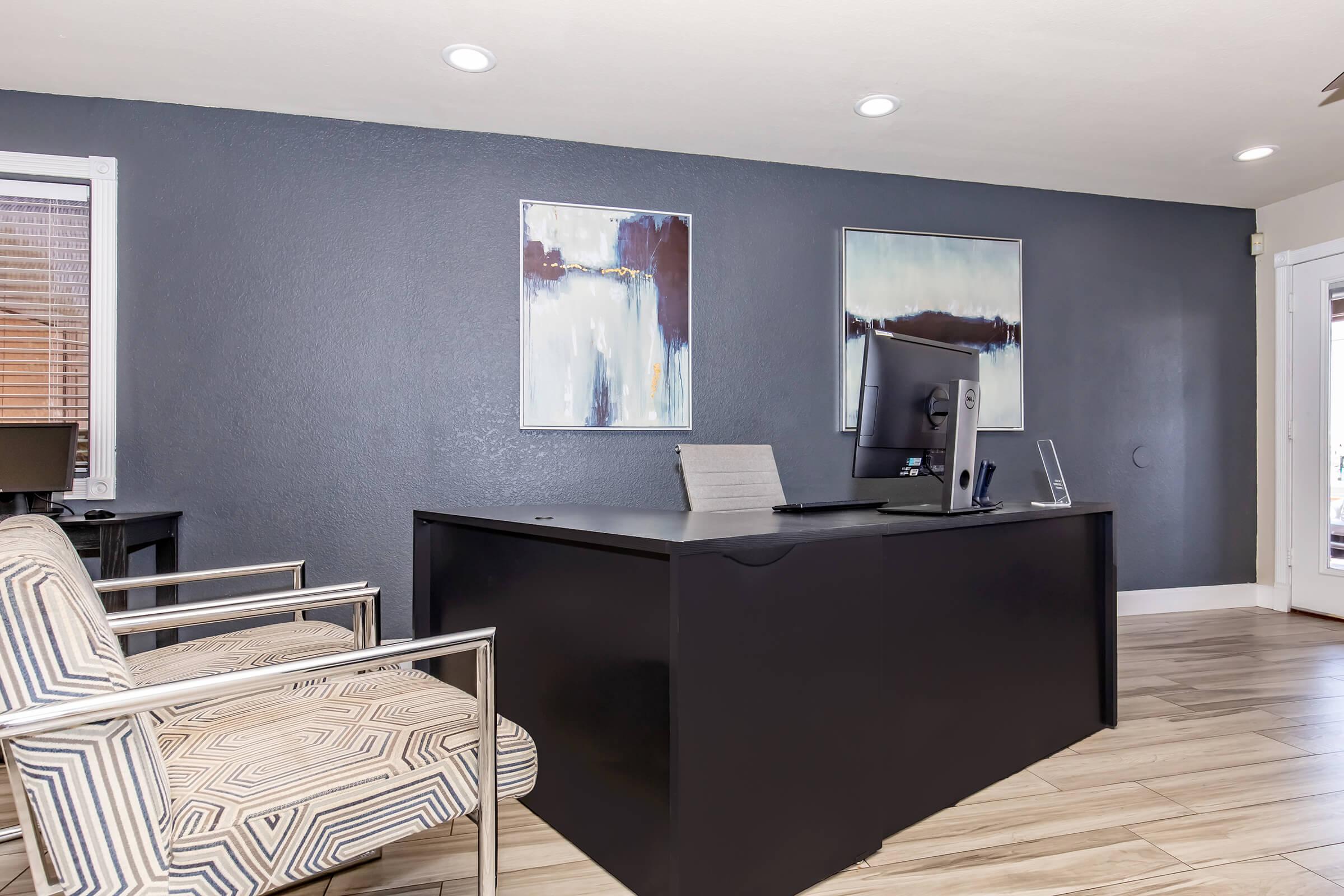 Modern office interior featuring a sleek black desk with a computer monitor, a gray and white patterned chair, and two abstract paintings on the wall. The room has neutral-colored walls and a doorway leading to an outdoor area, with ample natural light illuminating the space.
