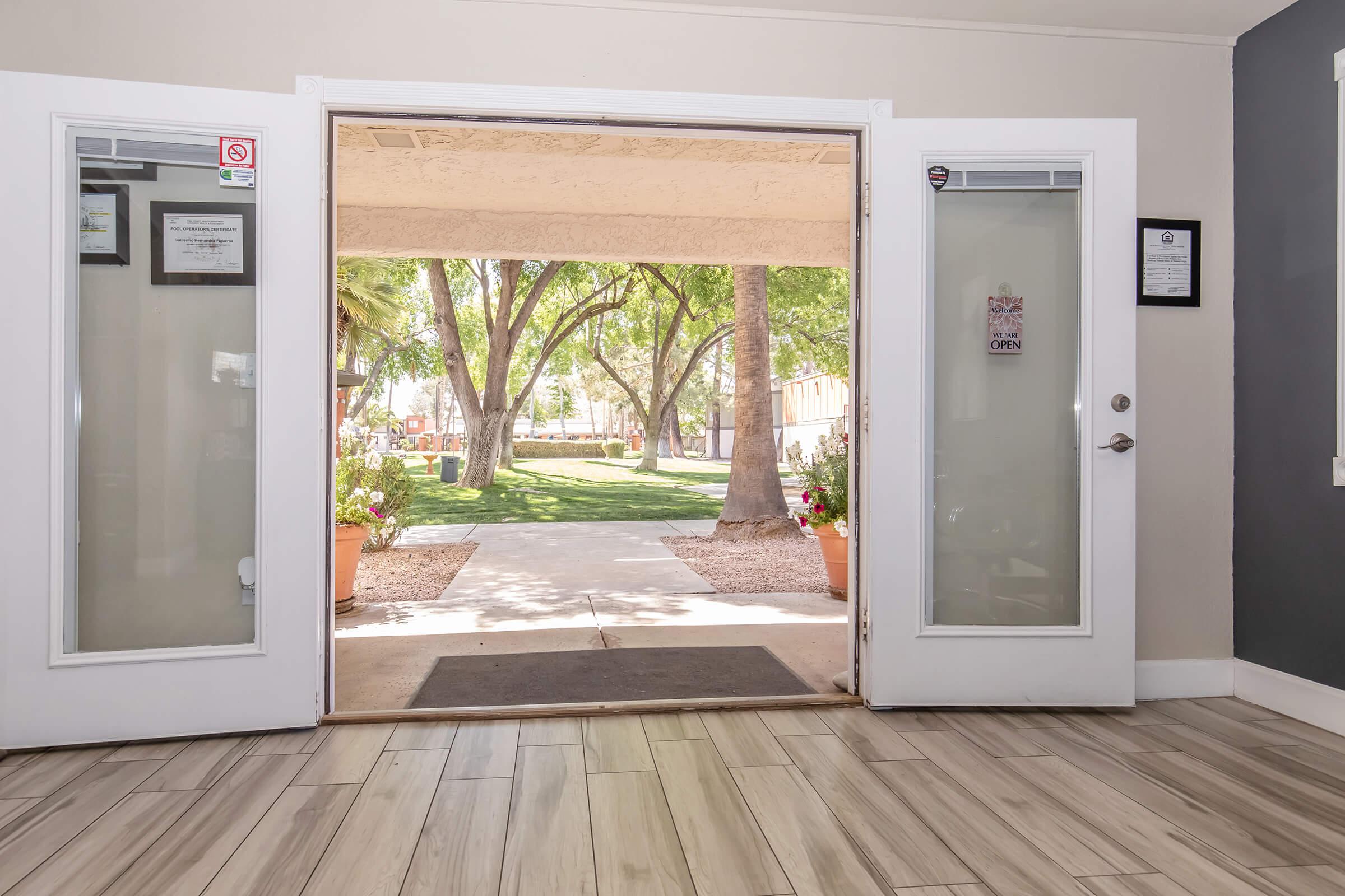View from the entrance of a building, showing a sunny pathway leading to a landscaped outdoor area with trees and plants. The glass doors are open, revealing a welcoming indoor space with neutral-colored walls and a wooden floor.