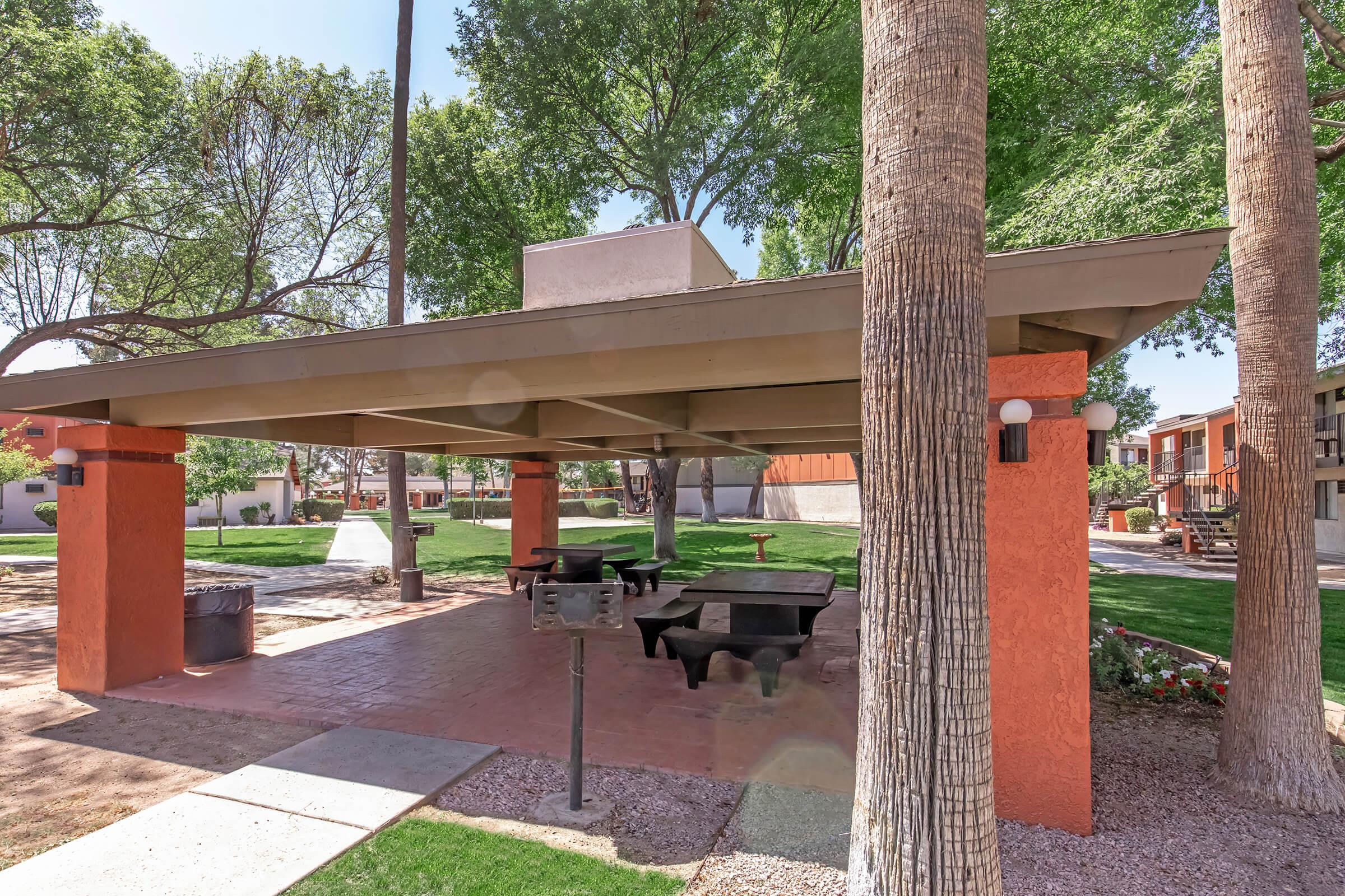 A shaded picnic area featuring a large pavilion with a flat roof supported by orange columns. The space includes wooden picnic tables and a grill, surrounded by lush green grass and tall trees. A trash can is visible nearby, with apartment buildings in the background.
