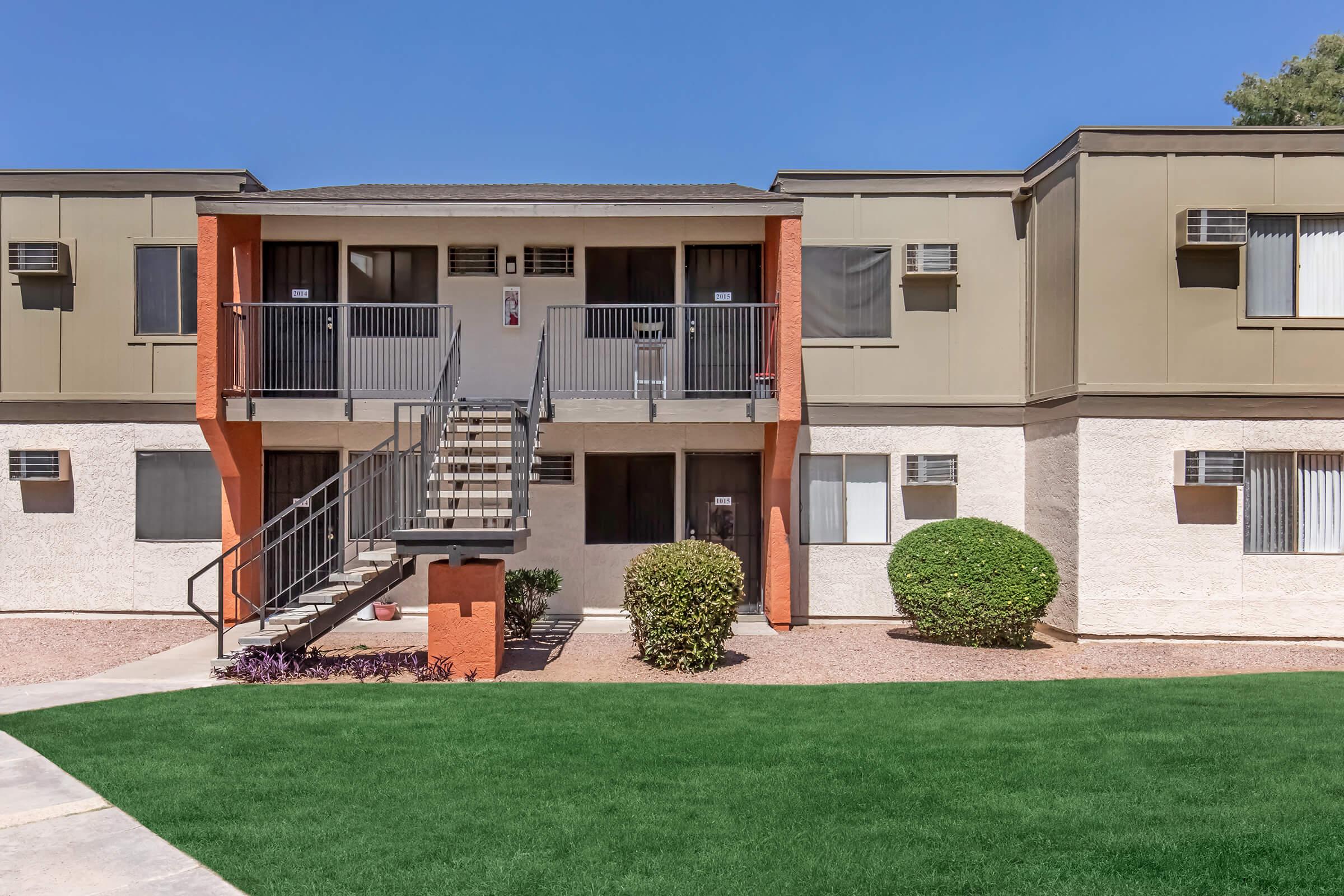Two-story apartment building with a mix of beige and orange exterior. A staircase leads to the second floor, with balcony railings visible. The ground is covered in green grass and decorative shrubs. Clear blue sky overhead. Air conditioning units are mounted on the walls.