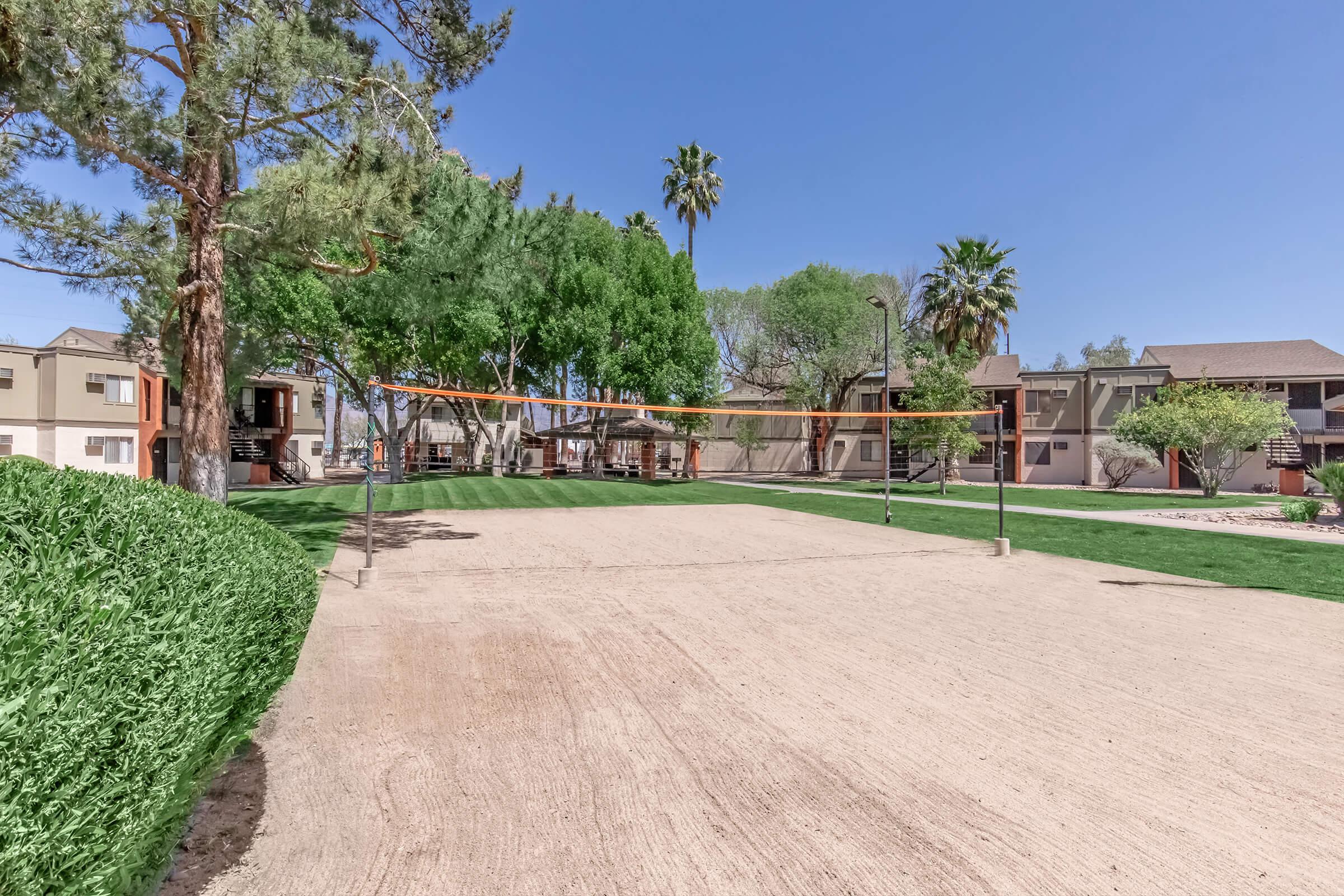 Volleyball court in a landscaped outdoor area surrounded by trees and palm trees, with two neighboring buildings in the background. The sandy court is ready for play, set against a clear blue sky.