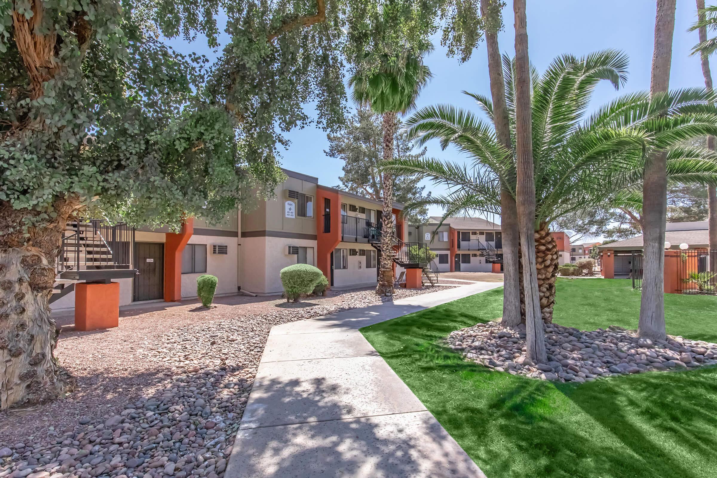 A landscaped pathway leads through a well-maintained apartment complex, featuring palm trees and shrubs. The building has a modern design with various colors and balconies visible. Green grass contrasts with the gravel and stone landscaping, creating a welcoming outdoor space.