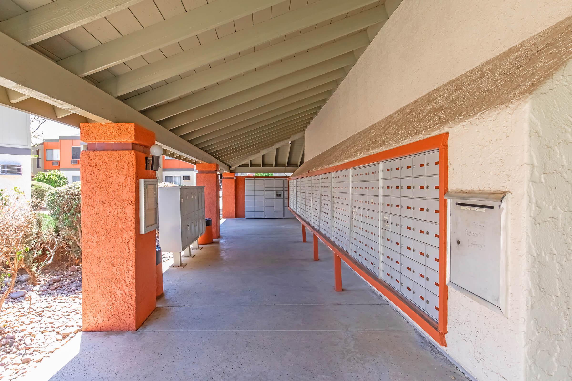 A covered mailbox area featuring multiple mailboxes on the left side, with a few bright orange pillars supporting the ceiling. The setting is clean and well-maintained, and there is a small mailbox on the right side. Lush greenery is visible outside the entrance.