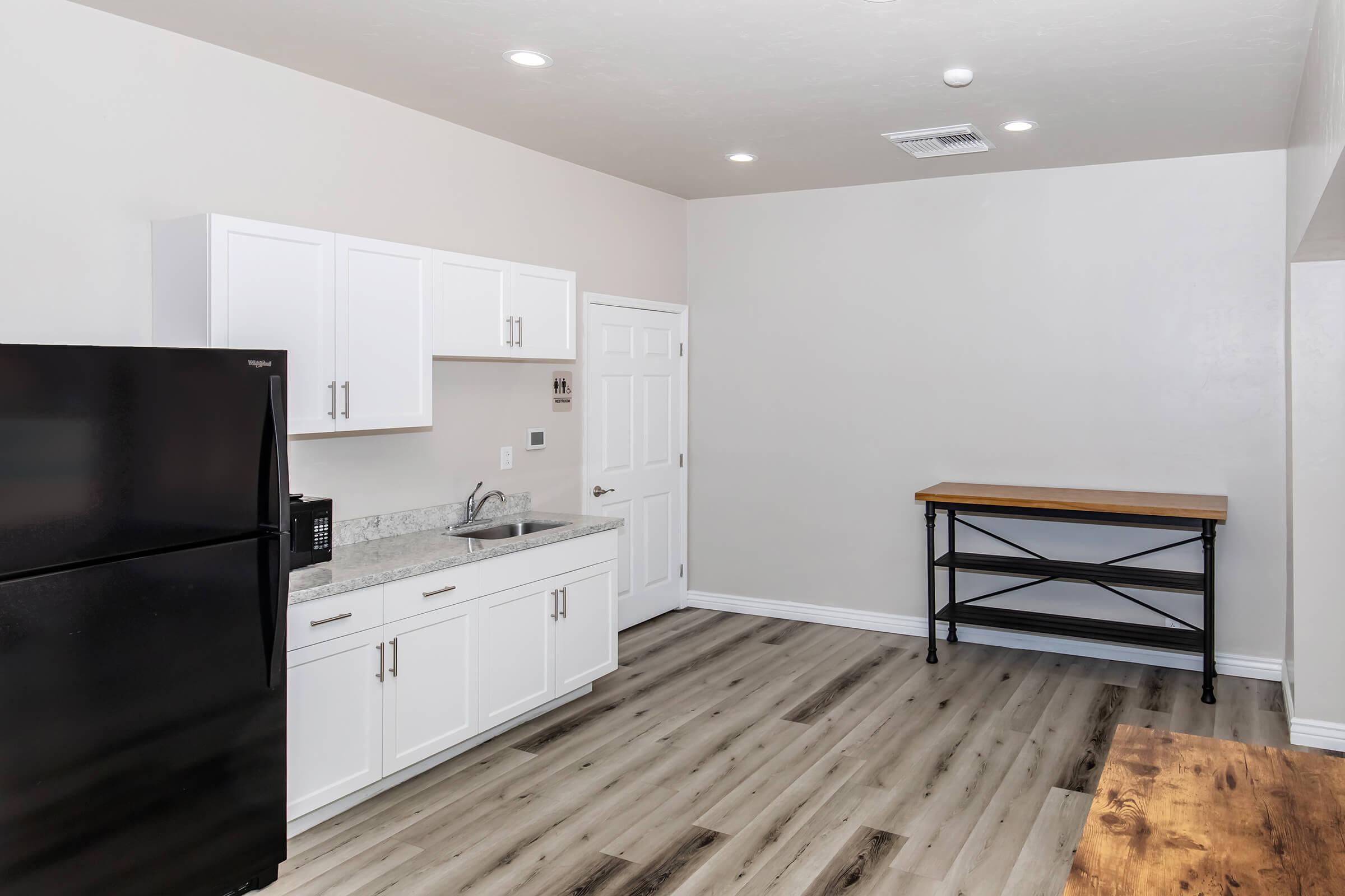 A modern kitchen featuring white cabinets, a black refrigerator, a microwave, and a countertop. The kitchen has light-colored walls and a wooden table with metal legs against one wall. The flooring is a light wood laminate, and there are overhead lights illuminating the space.