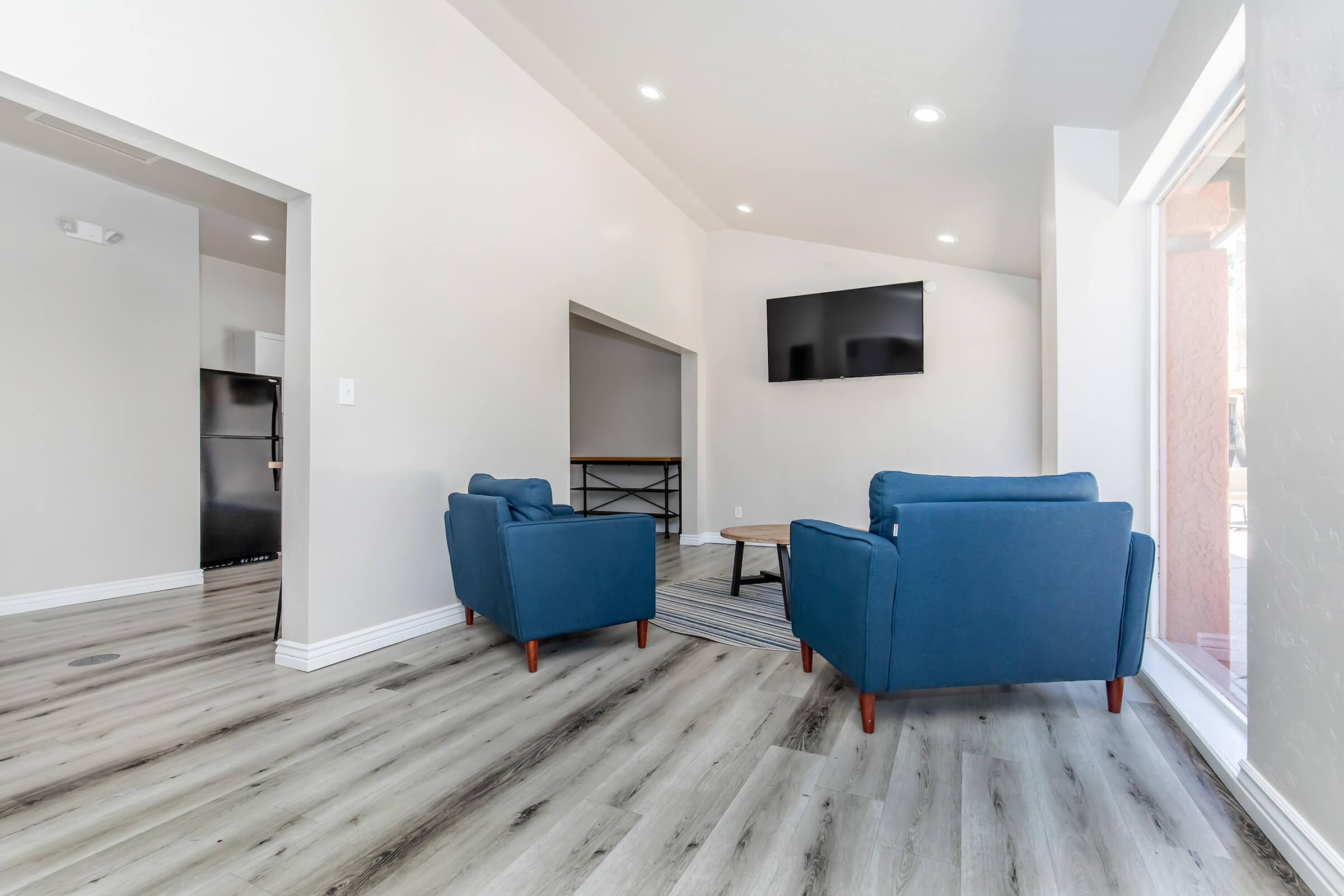 A modern living area featuring two blue armchairs and a wooden coffee table on a light-colored floor. A flat-screen TV is mounted on the wall, and large windows provide natural light. The space is stylishly decorated with neutral tones and minimalistic design.