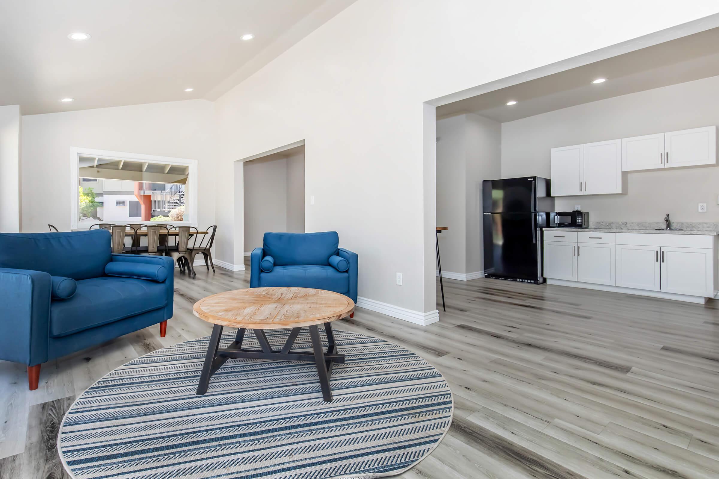 A modern, open living space featuring two blue armchairs, a round wooden coffee table on a striped rug, and a kitchenette with white cabinets and a black refrigerator. Natural light streams in, enhancing the airy feel of the room, with a dining area visible in the background.