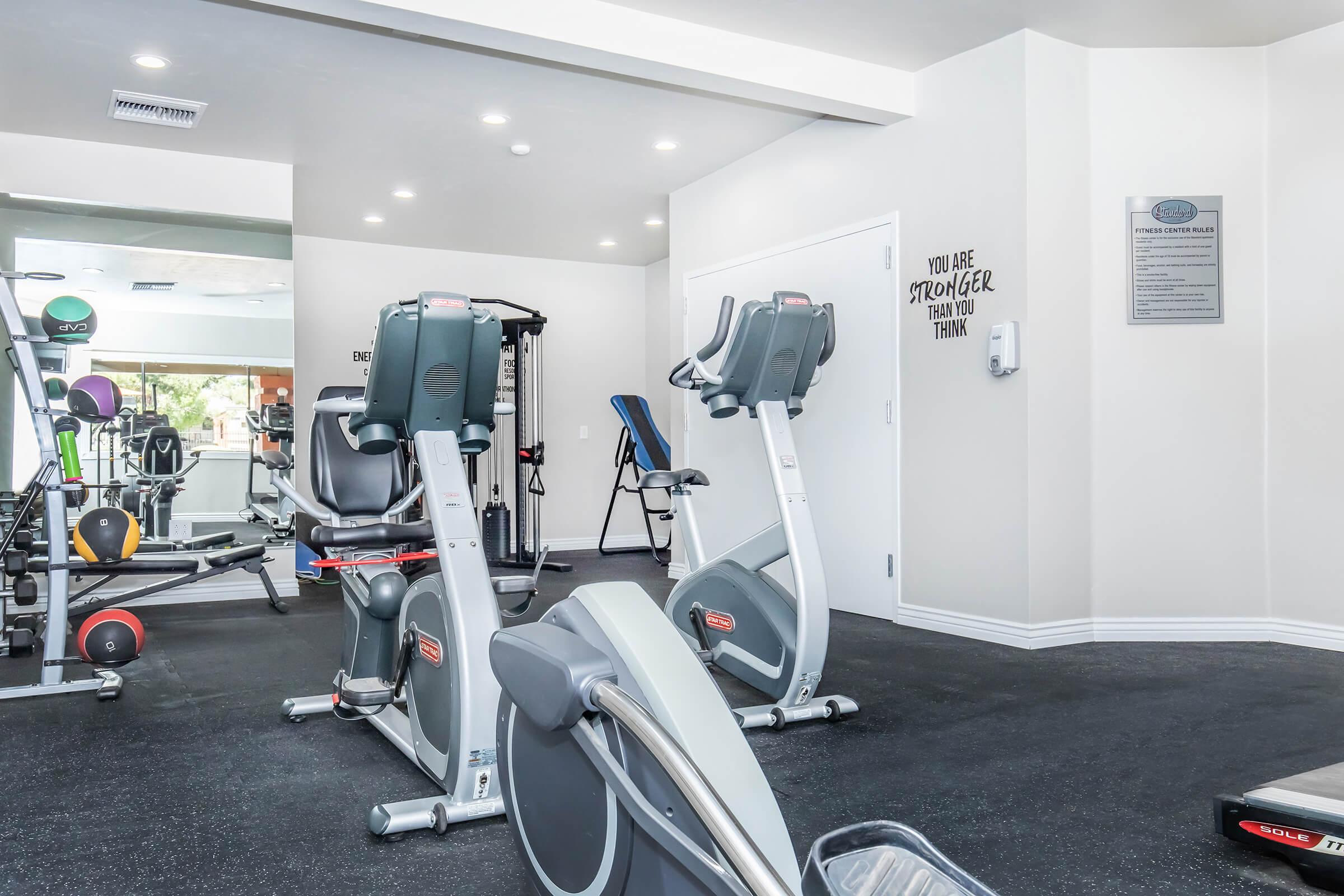 A modern gym space featuring exercise equipment, including stationary bikes and free weights. The walls are decorated with motivational quotes. The floor is covered in black rubber matting, and there is a large mirror reflecting the workout area. Bright, well-lit environment for fitness activities.