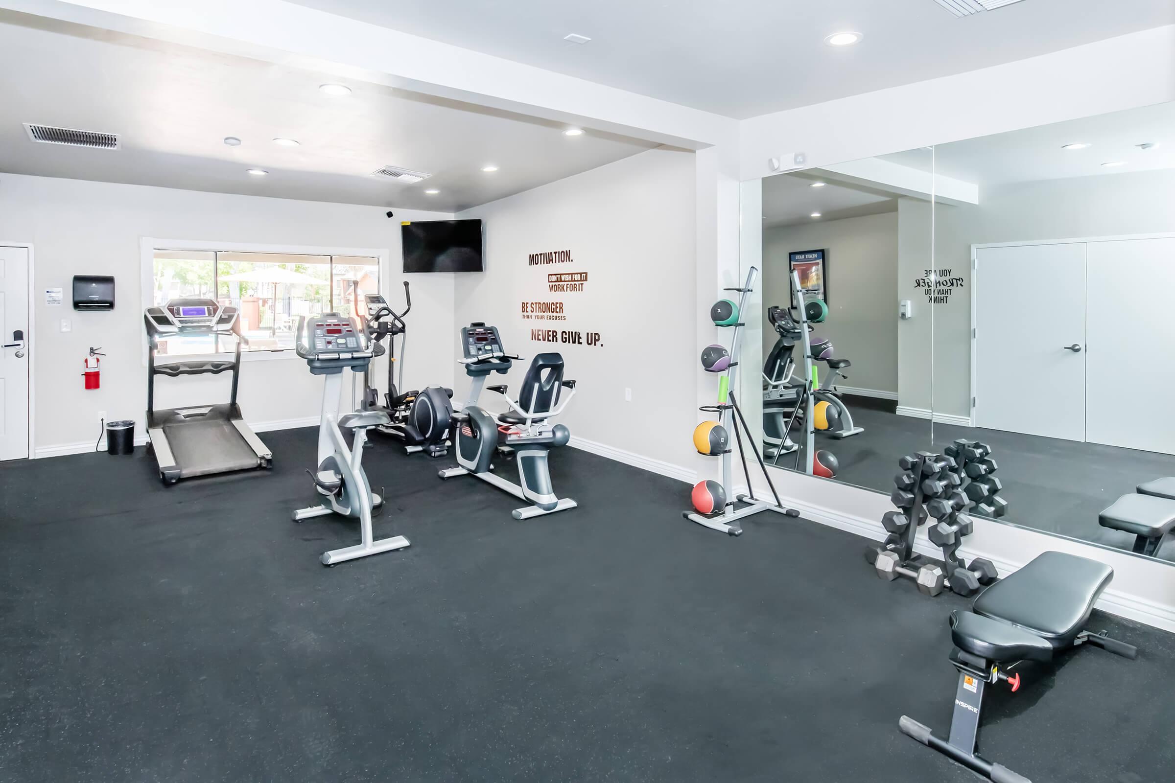 A bright and modern home gym featuring cardio equipment like a treadmill, weights, and a mirror. The wall displays a motivational quote, "Never give up." The flooring is black, creating a clean and sleek atmosphere for workouts.
