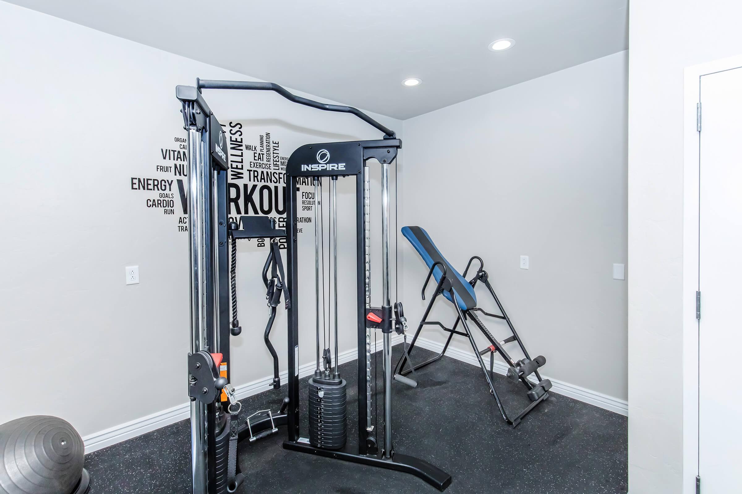 A modern home gym featuring a multi-gym machine, an inversion table, and fitness motivational wall art. The space has a neutral-colored wall and a black floor mat, creating a sleek and organized workout environment.