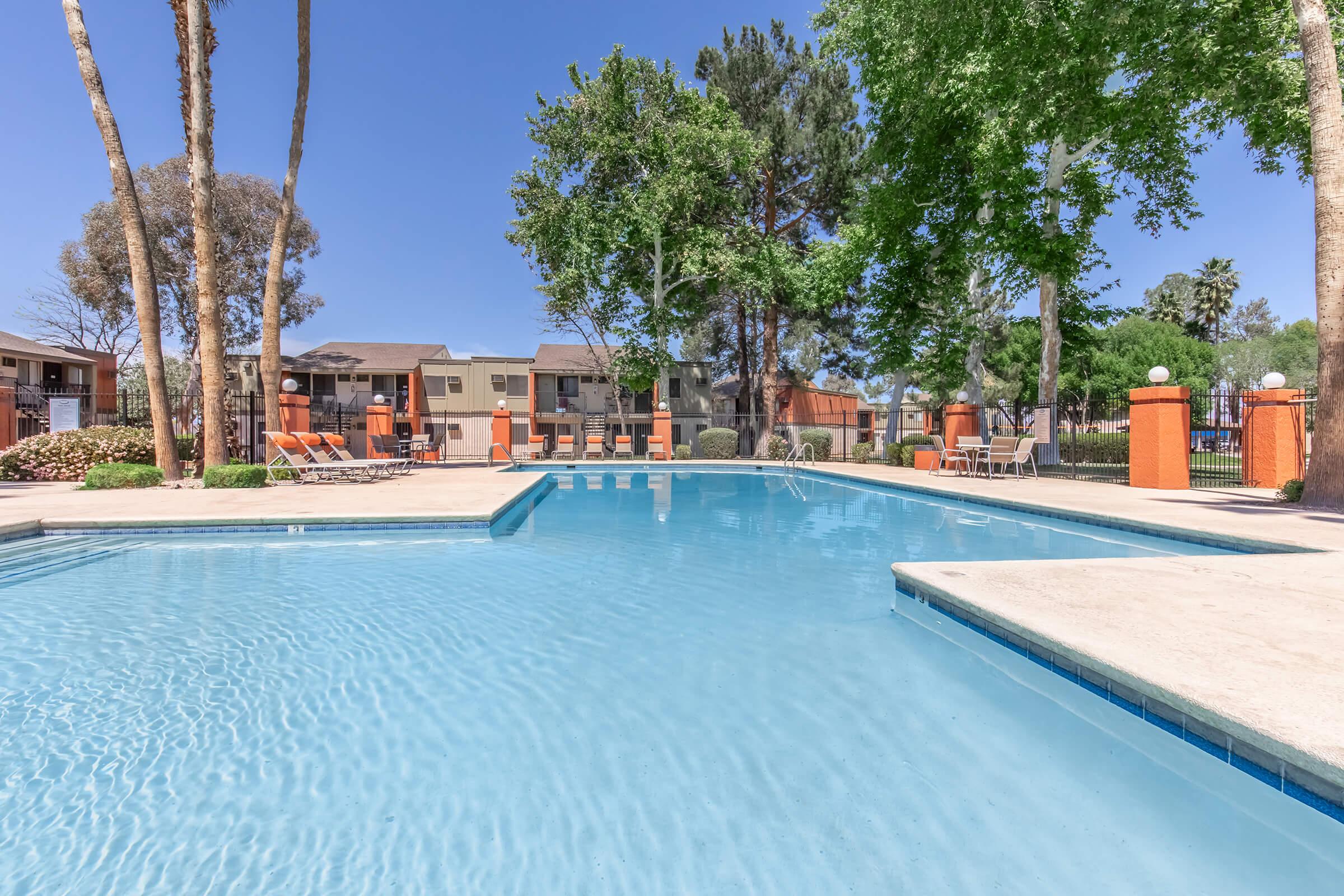 A clear swimming pool surrounded by lush greenery and tall trees. Lounge chairs are arranged near the pool, and several buildings are visible in the background, showcasing a residential area. The sky is bright blue, indicating a sunny day.