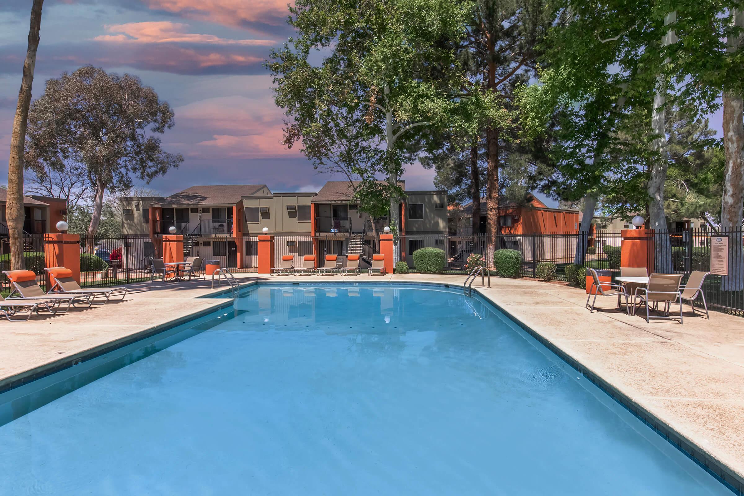 A clear blue swimming pool surrounded by lounge chairs and trees. Several apartments are visible in the background, with an orange and brown color scheme. The sky features soft pink and purple hues, suggesting sunrise or sunset. The scene is tranquil and inviting, perfect for relaxation.