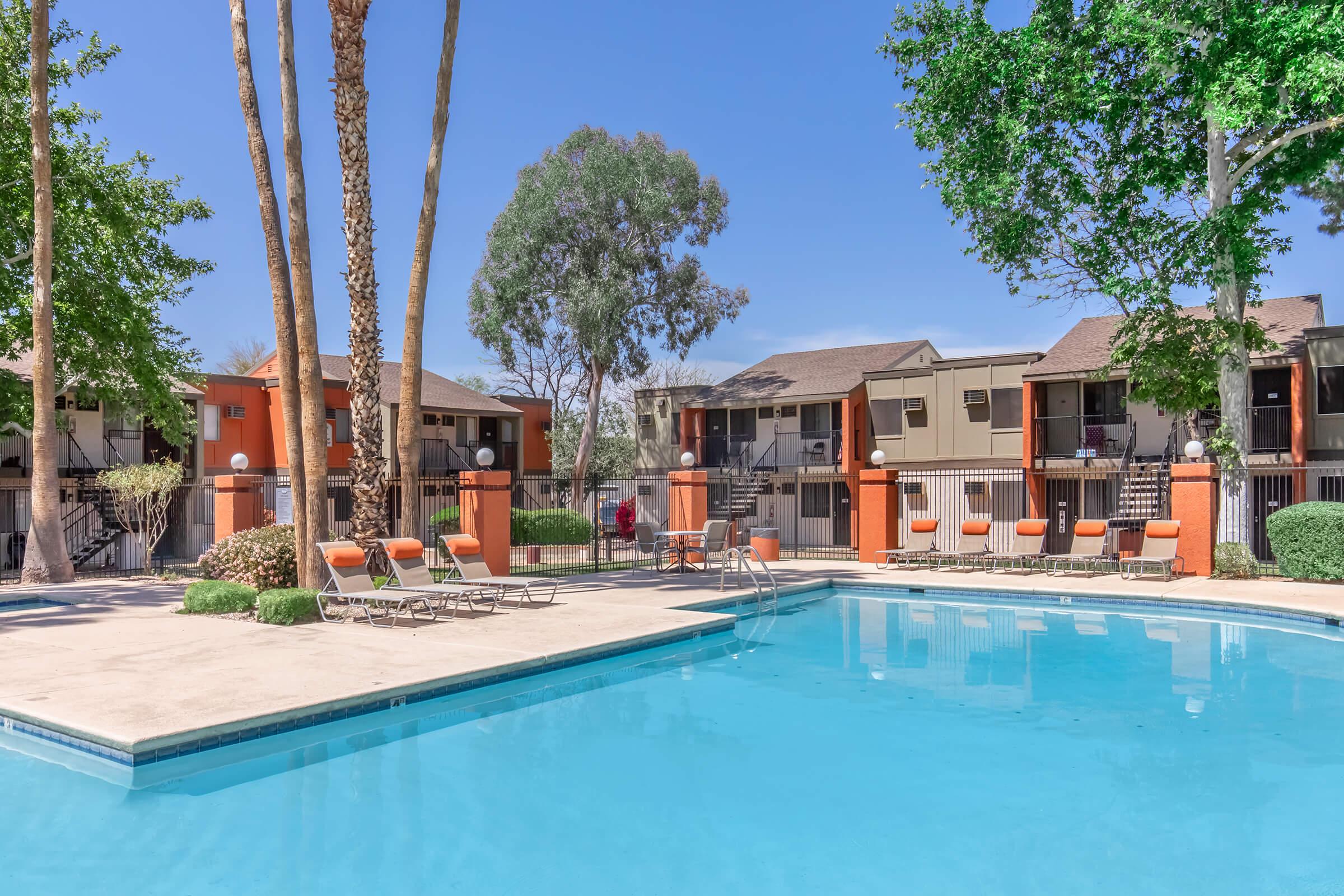 A bright outdoor swimming pool surrounded by palm trees and lounge chairs, with orange accents. In the background, there are buildings with balconies, and clear blue skies above. The area appears inviting and well-maintained, perfect for relaxation and leisure.